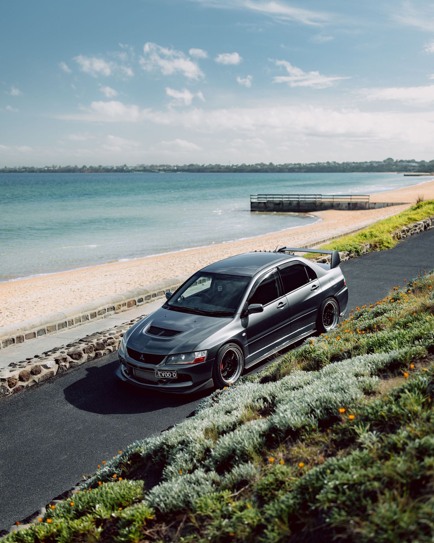 A dark grey Mitsubishi Lancer Evolution parked on a coastal road with a sandy beach and calm blue ocean in the background.