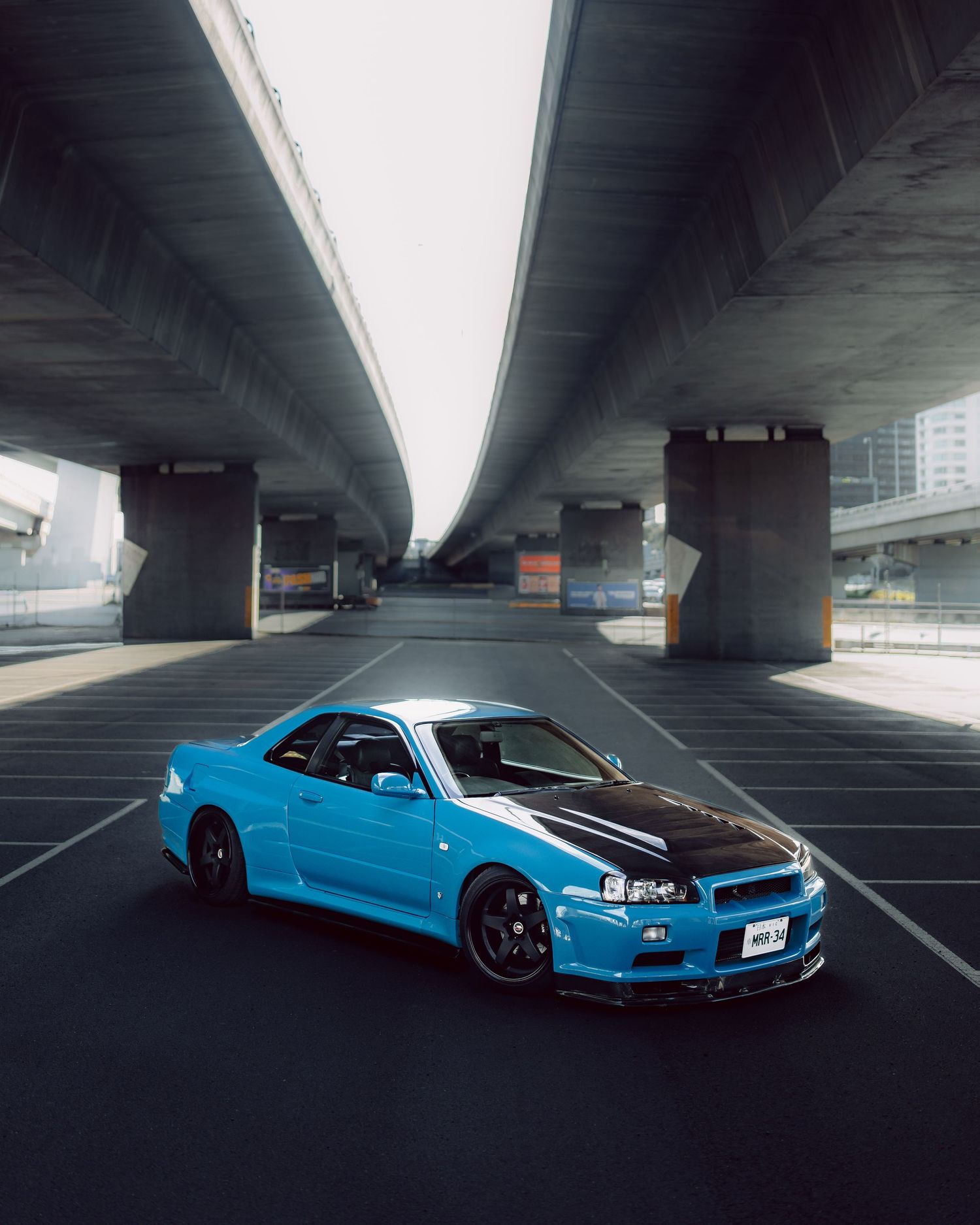 A blue Nissan Skyline GT-R R34 parked under concrete highway overpasses on an empty lot.