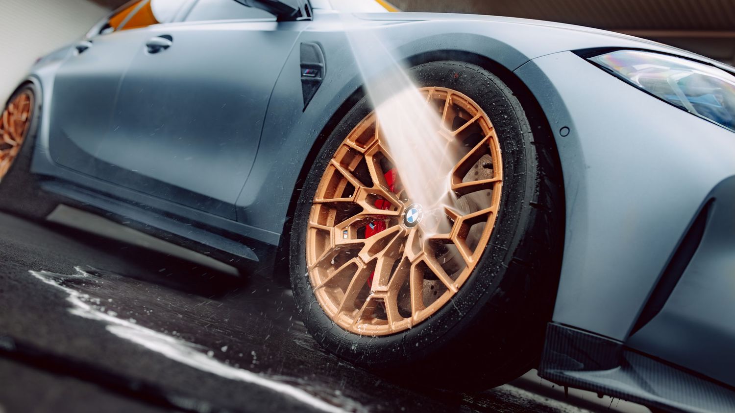 A blue sports car with gold alloy wheels being washed with a high-pressure water jet at a car wash.