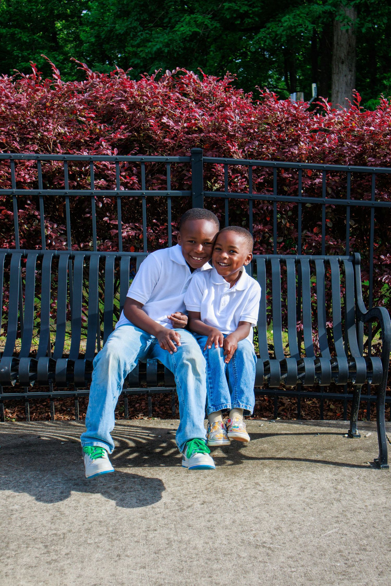 Two young boys in white shirts and jeans sitting together on a black park bench with red bushes behind them.