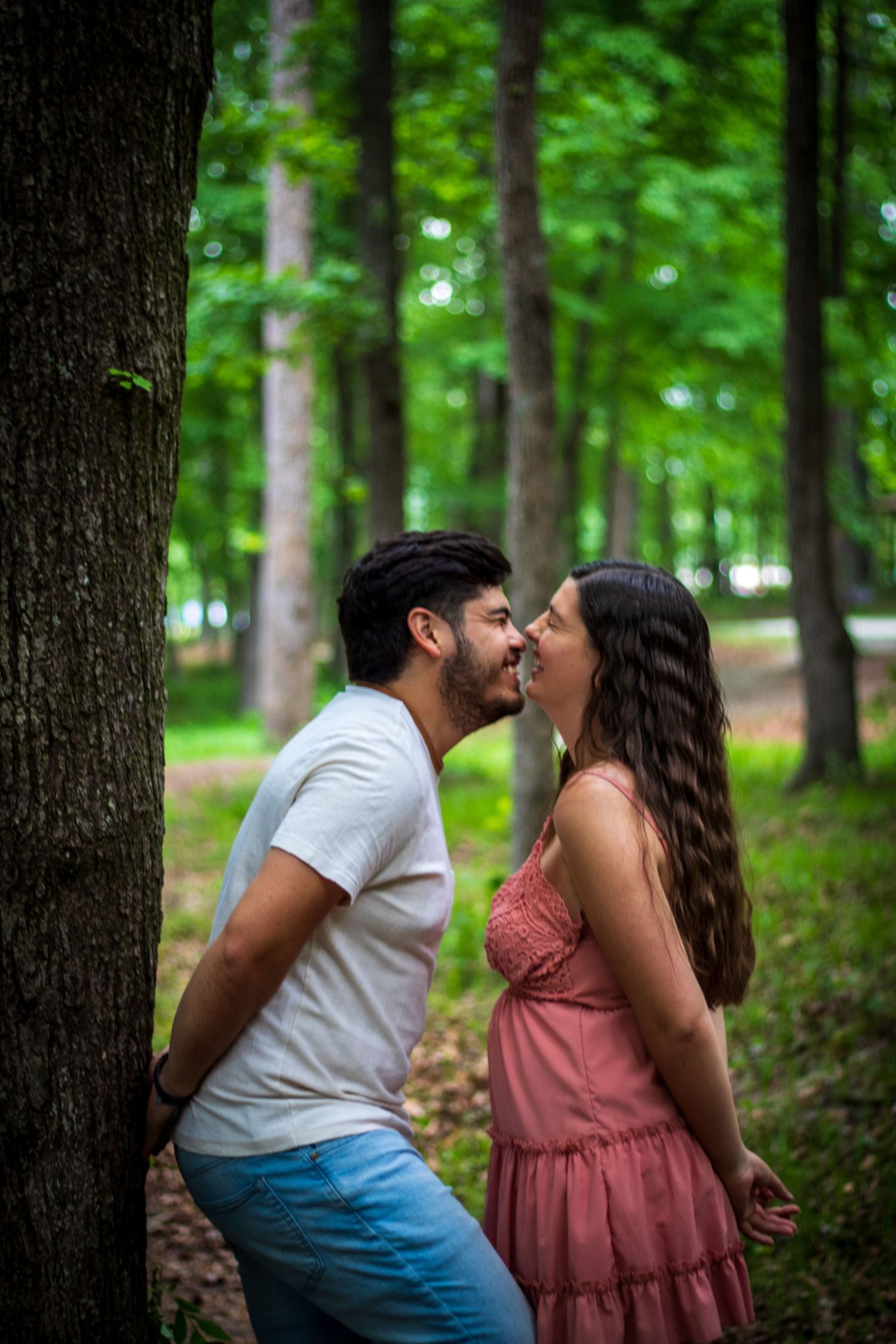 A romantic couple leans in for a kiss beside a large tree trunk in a lush green forest.