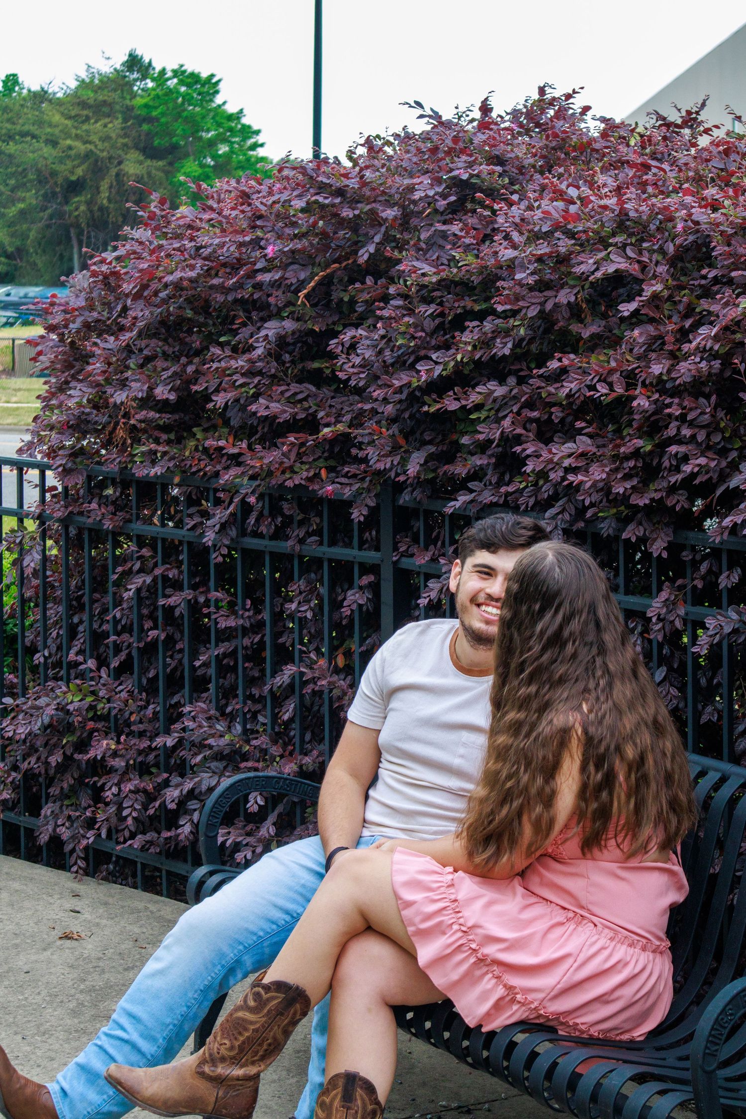 A smiling couple sits on a bench surrounded by purple foliage, the woman in a pink dress leaning close to the man in a white tee.