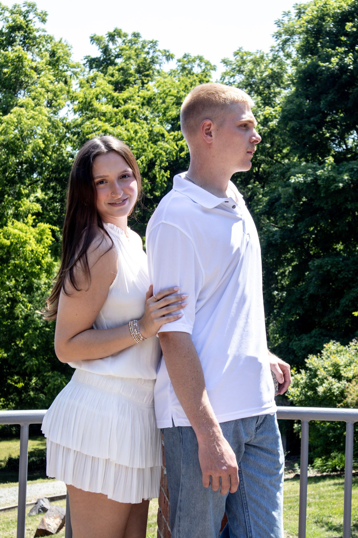 Young couple in white outfits posing together outdoors on a sunny day with lush green trees in the background.
