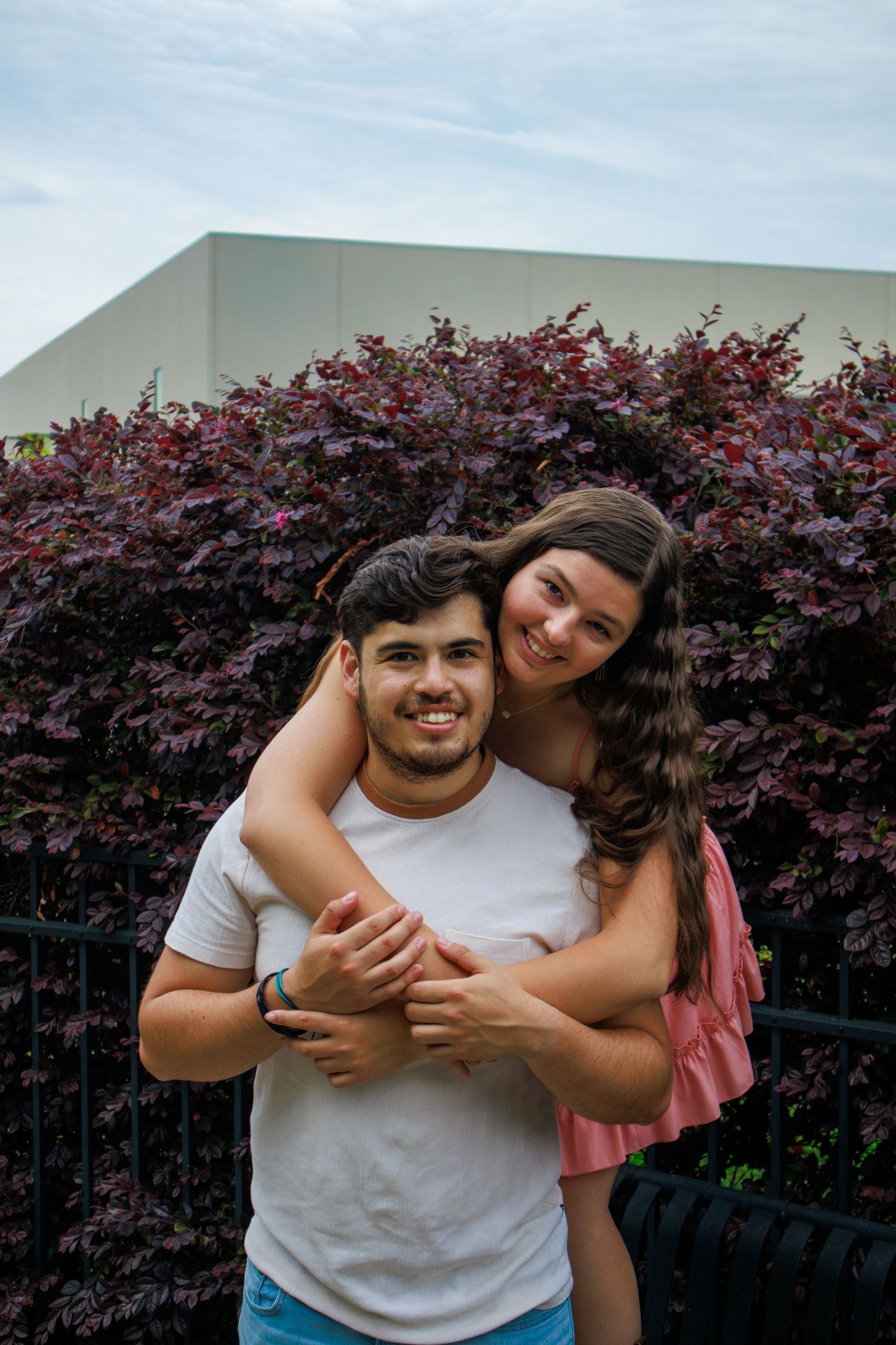 Couple embracing outdoors, woman hugging man from behind, both smiling in front of purple bushes.