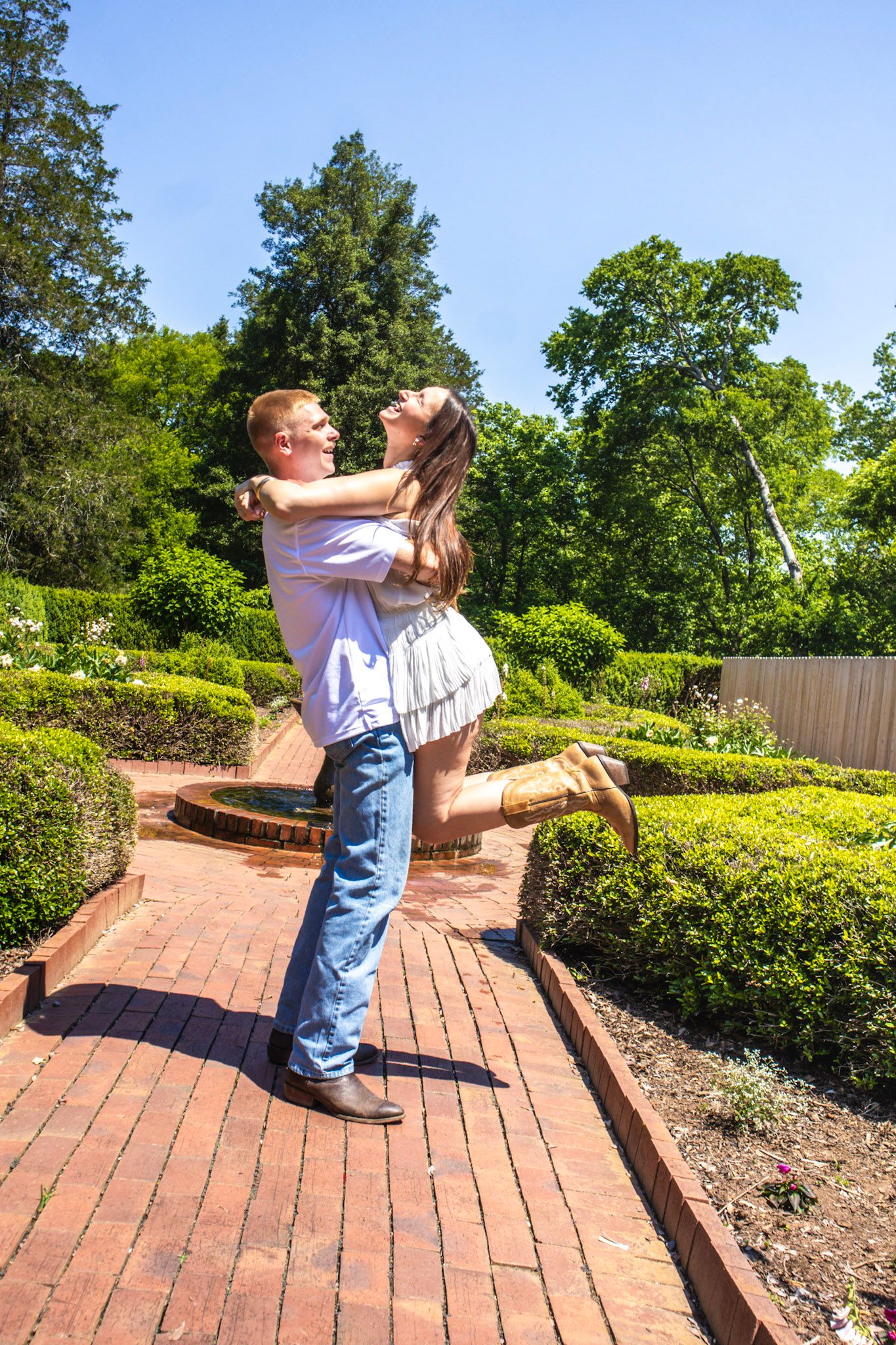 Couple embracing and lifting in a lush garden with brick pathways, green hedges, and trees under a clear blue sky.