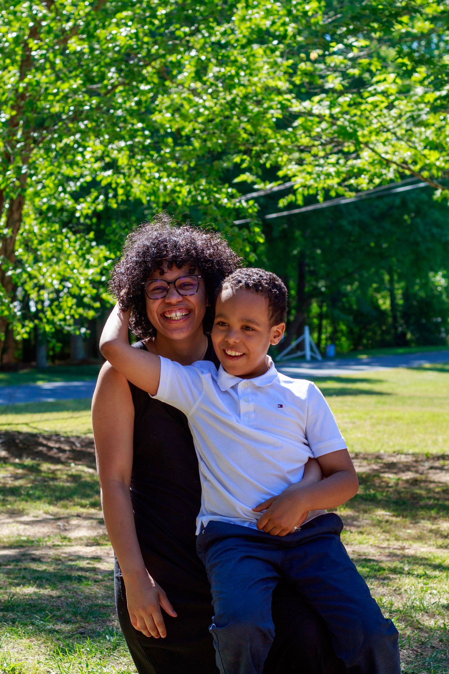 A smiling woman with curly hair and glasses holds a young boy in a white polo shirt outdoors in a sunny park.