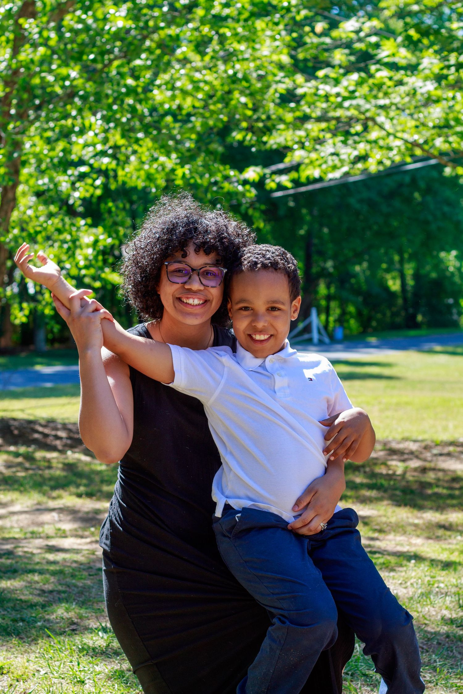 A smiling woman with glasses holds a young boy outdoors in a lush green park on a sunny day.