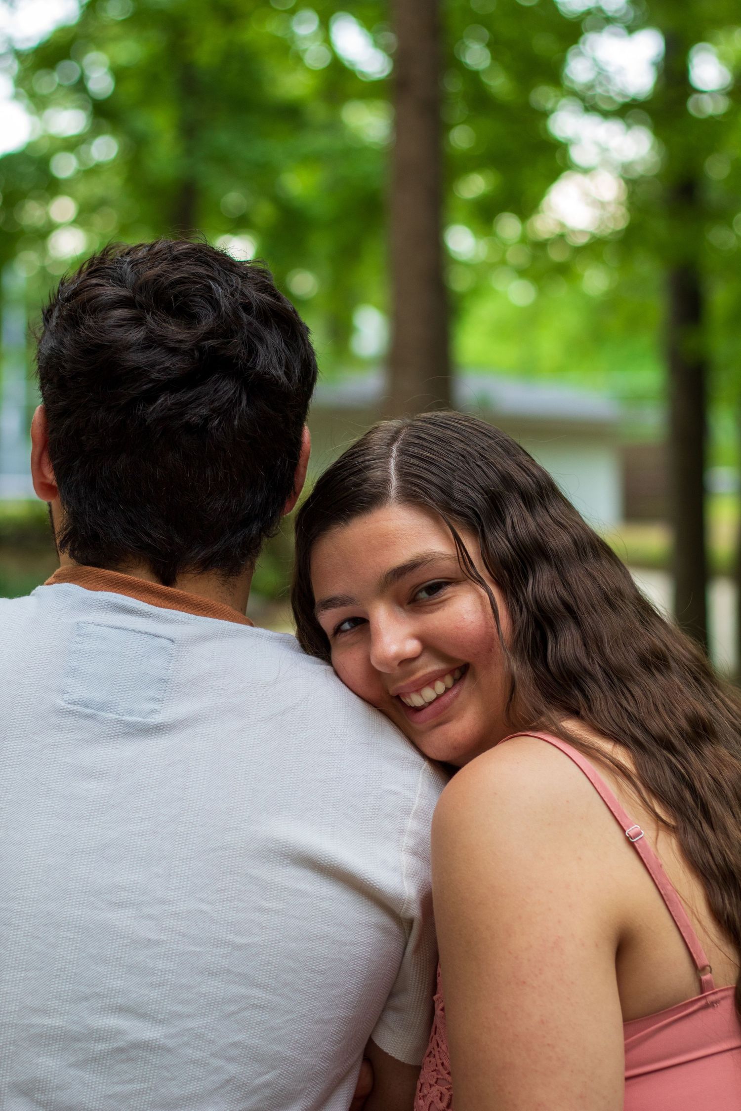 A smiling young woman with long hair hugs a man from behind outdoors, surrounded by lush green trees.