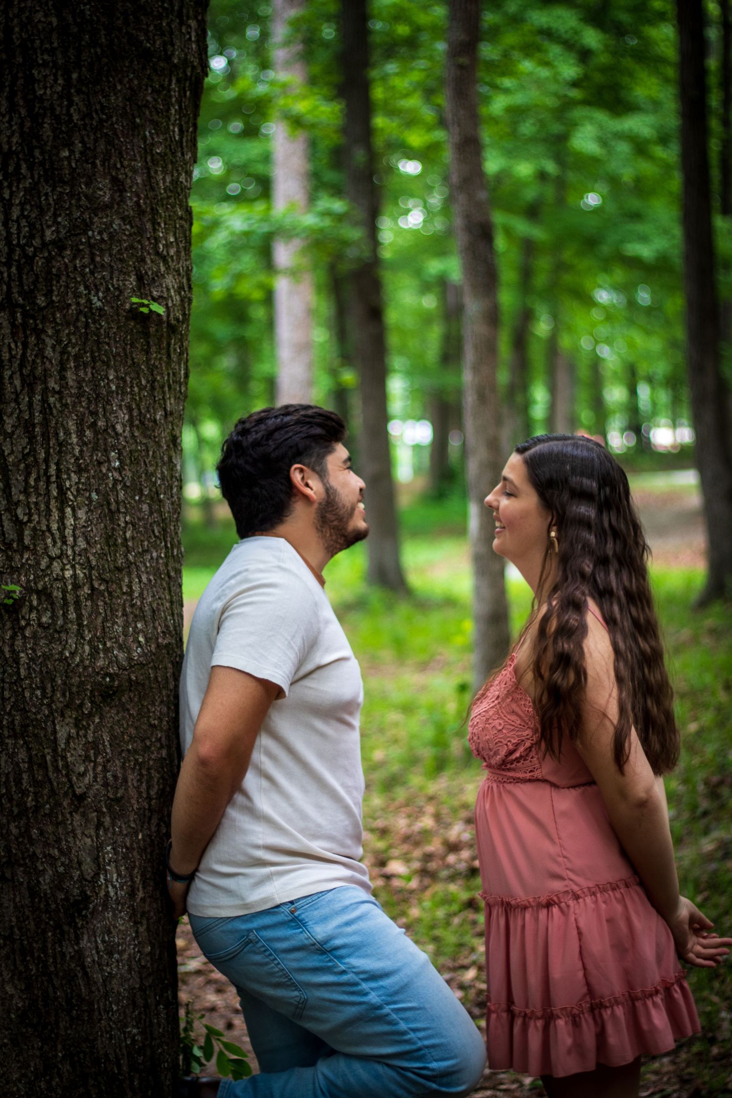 A couple smiles at each other while standing by a tree trunk in a sunlit green forest.