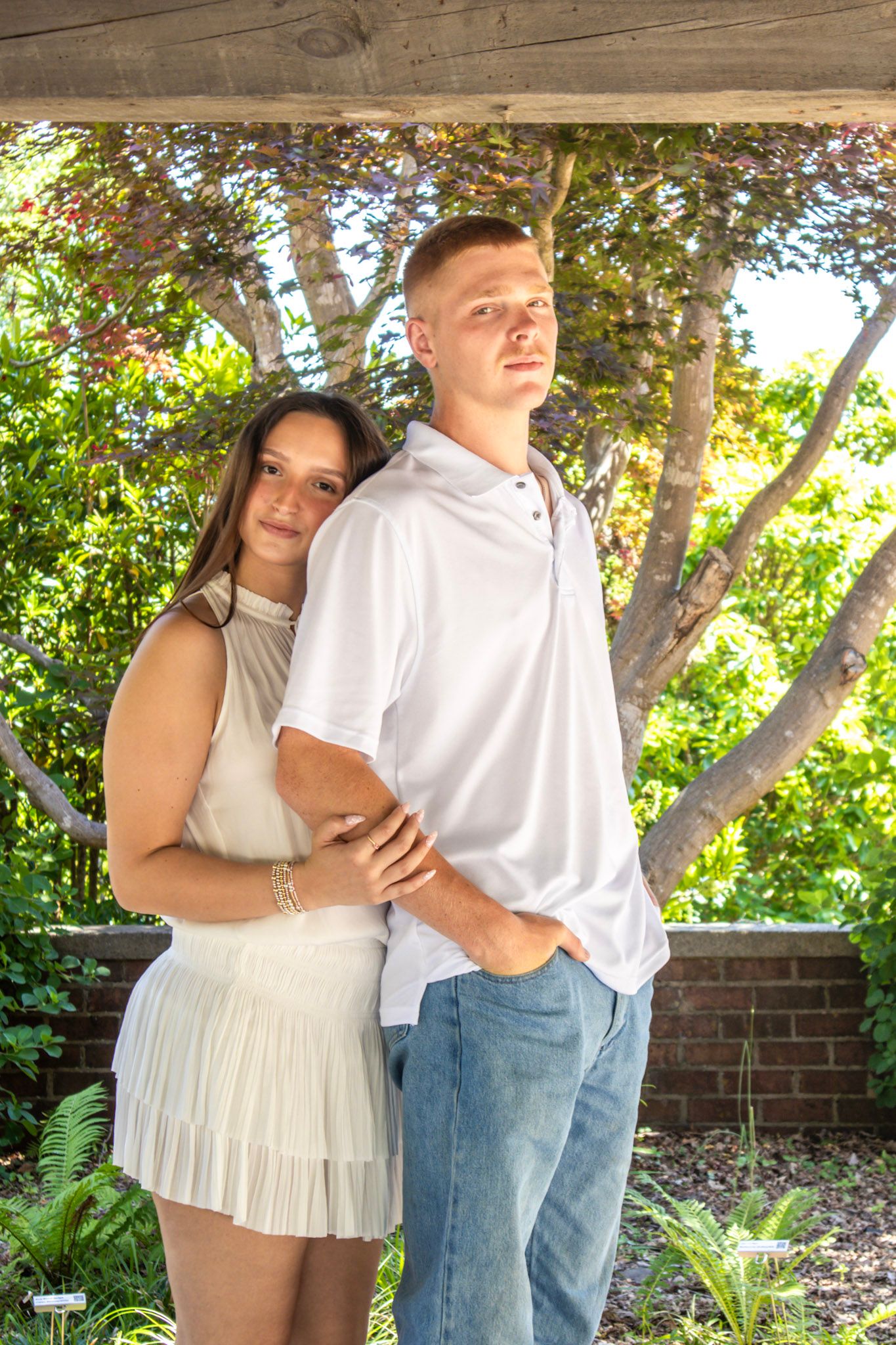 A young couple poses outdoors under a wooden pergola surrounded by lush green trees, wearing white outfits.