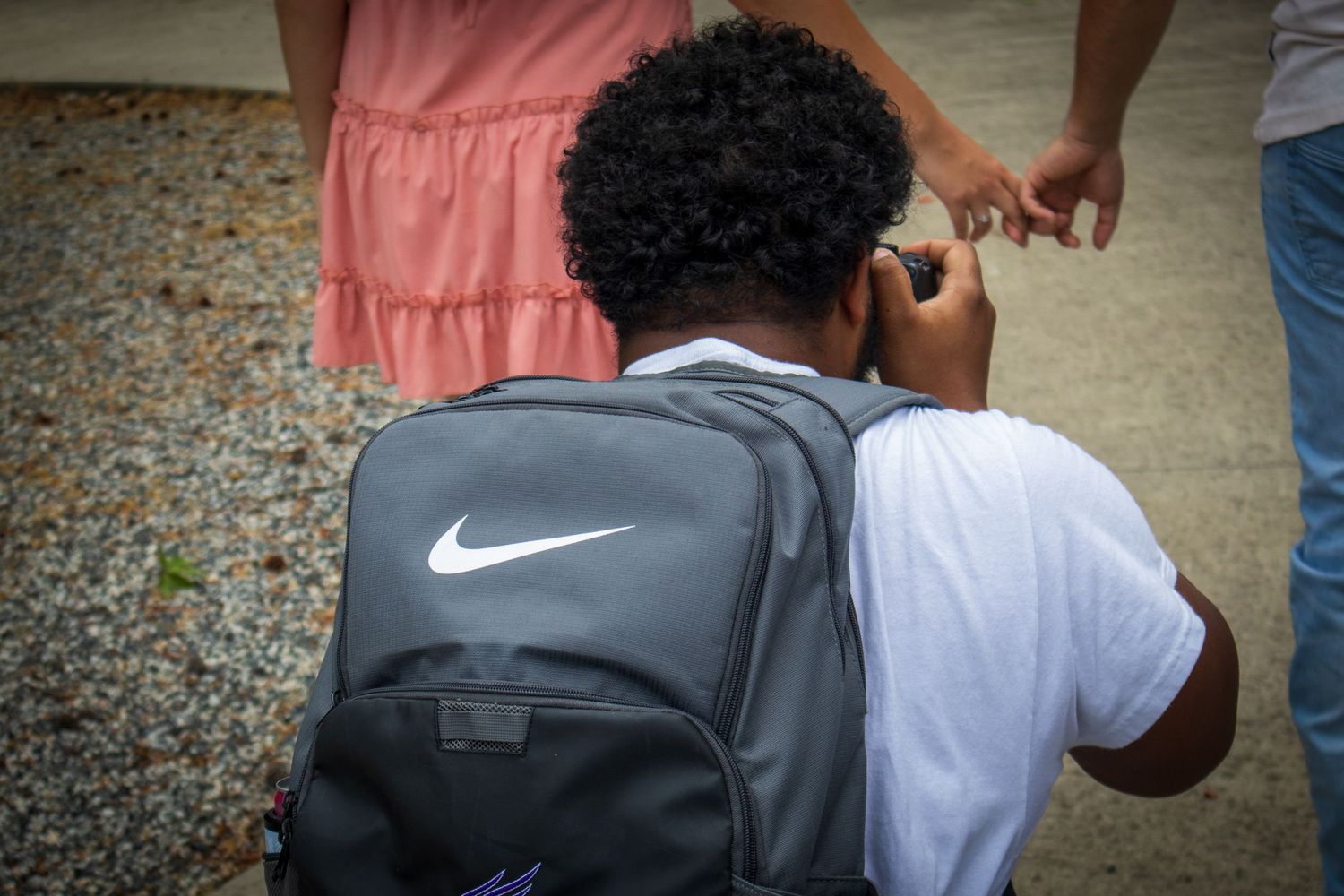 A child wearing a gray Nike backpack stands on gravel next to a person in a pink dress outdoors.