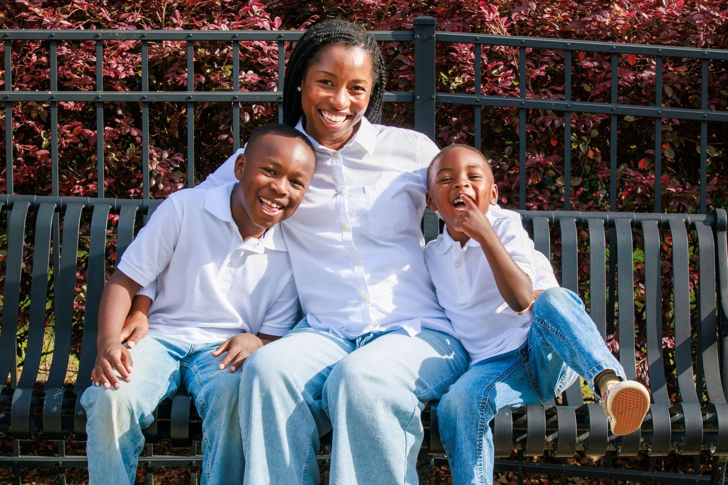A joyful African American family of three smiling together on a park bench, wearing matching white shirts and jeans.
