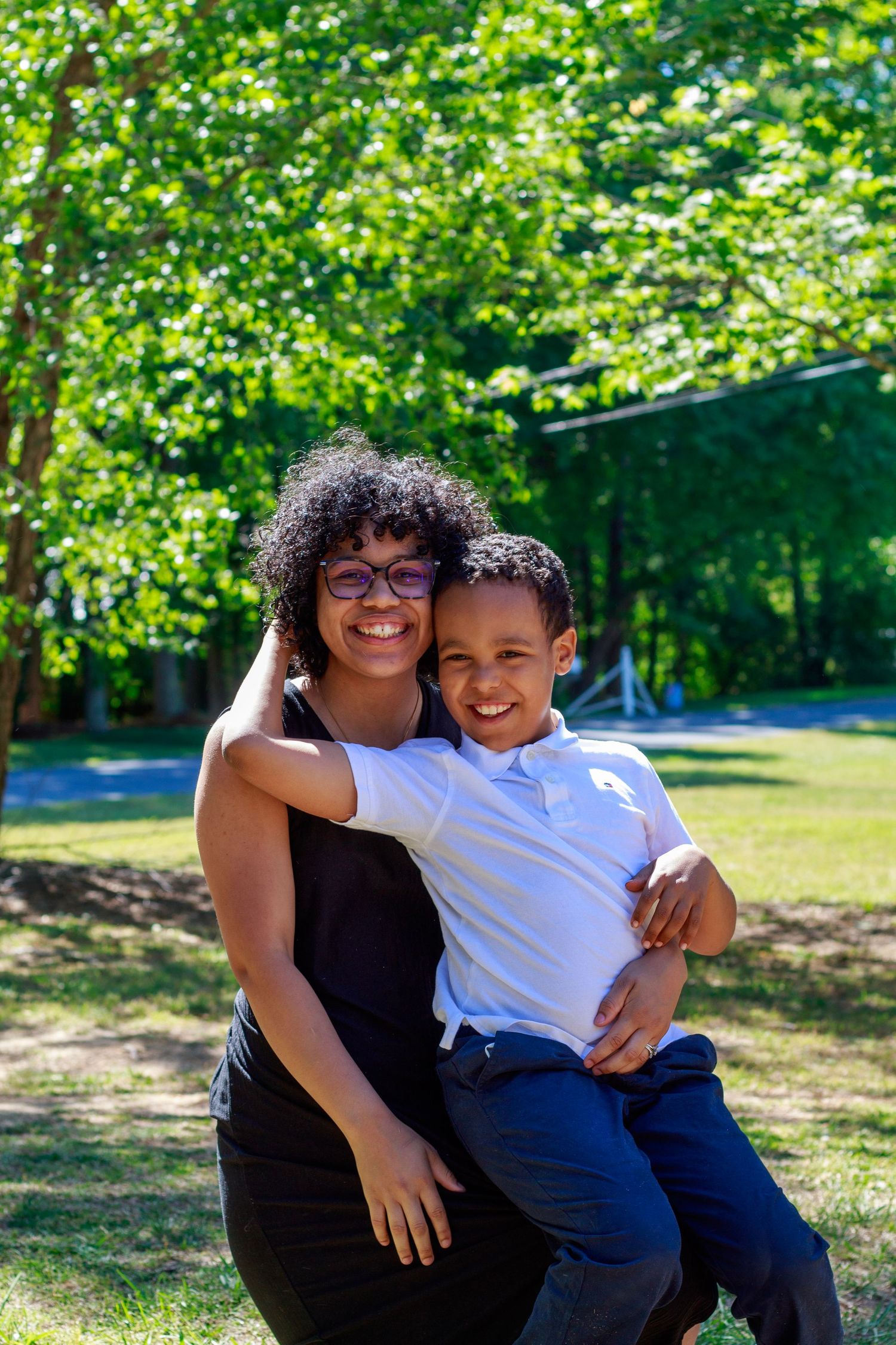 A woman with curly hair and glasses smiles while holding a young boy outdoors in a sunny green park.