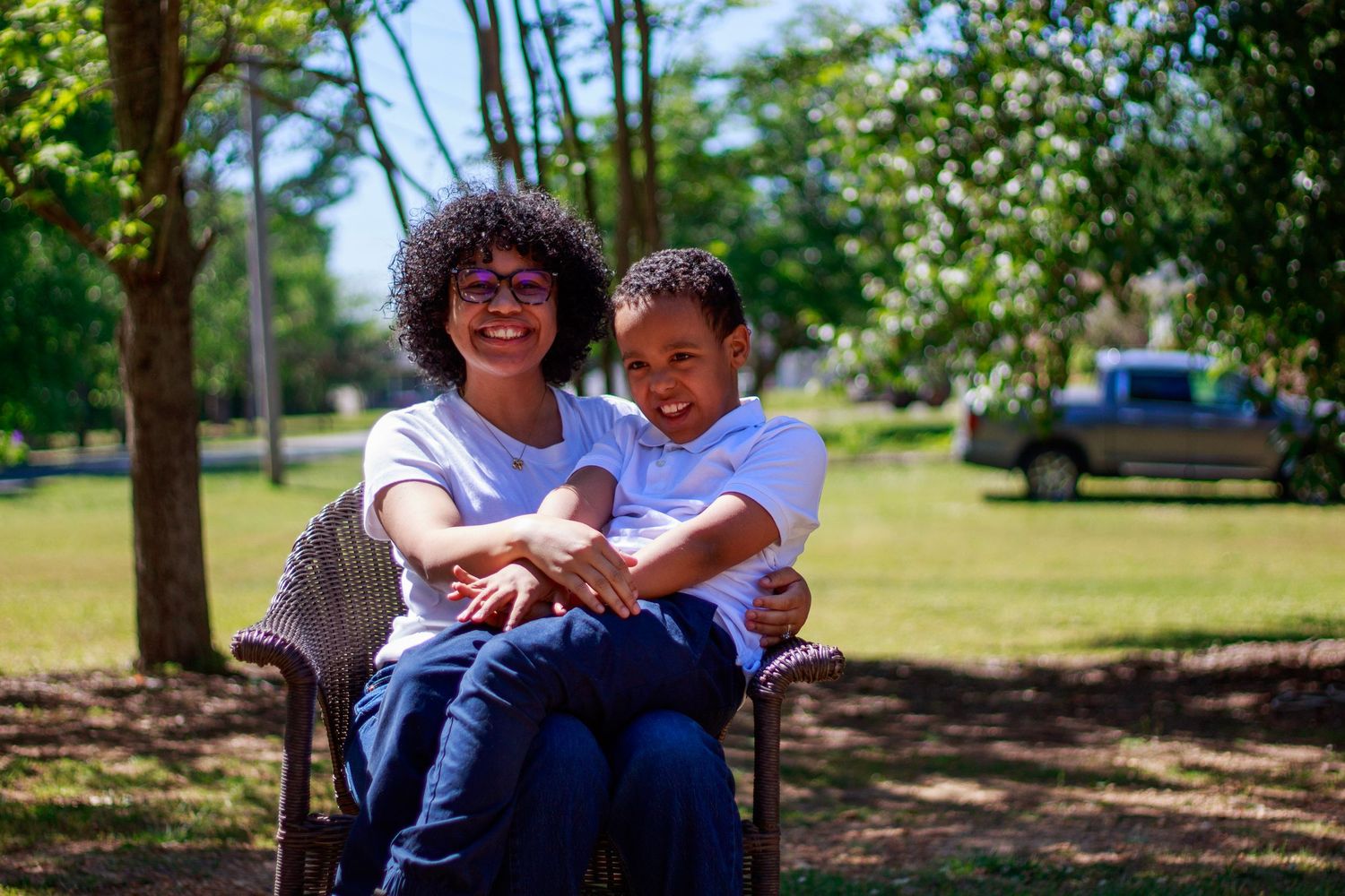 A woman and young boy smile together on a bench in a sunny green park.