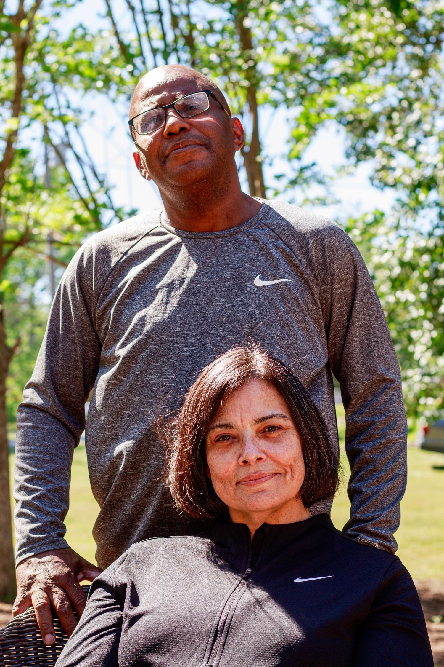 A man and woman wearing Nike athletic wear pose together outdoors among green trees on a sunny day.