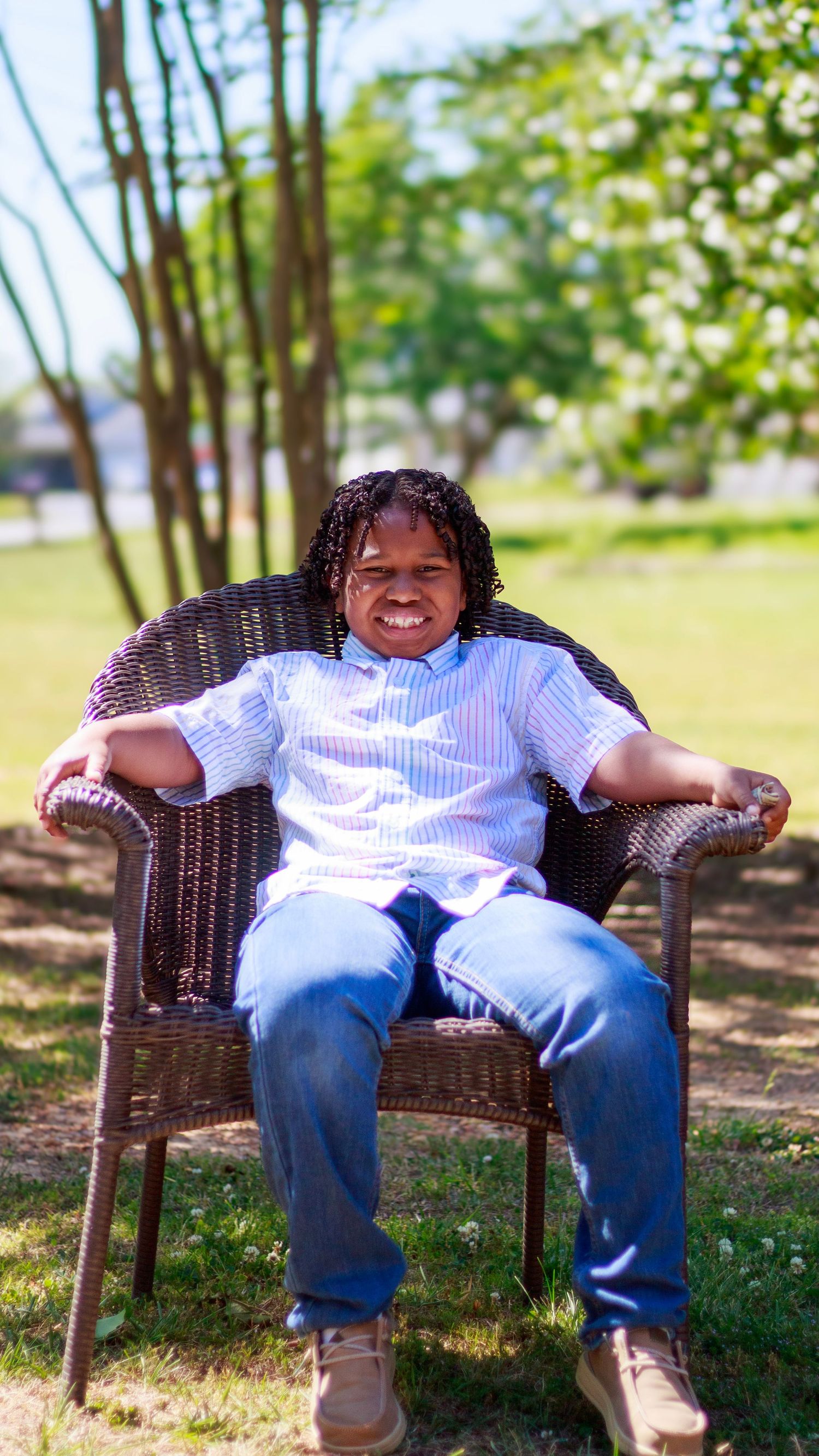 Smiling person with braids sitting relaxed in wicker chair outdoors, wearing striped shirt and jeans on sunny day.