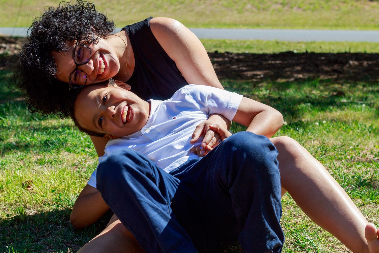 A woman and child laughing and playing together on green grass in a sunny park.
