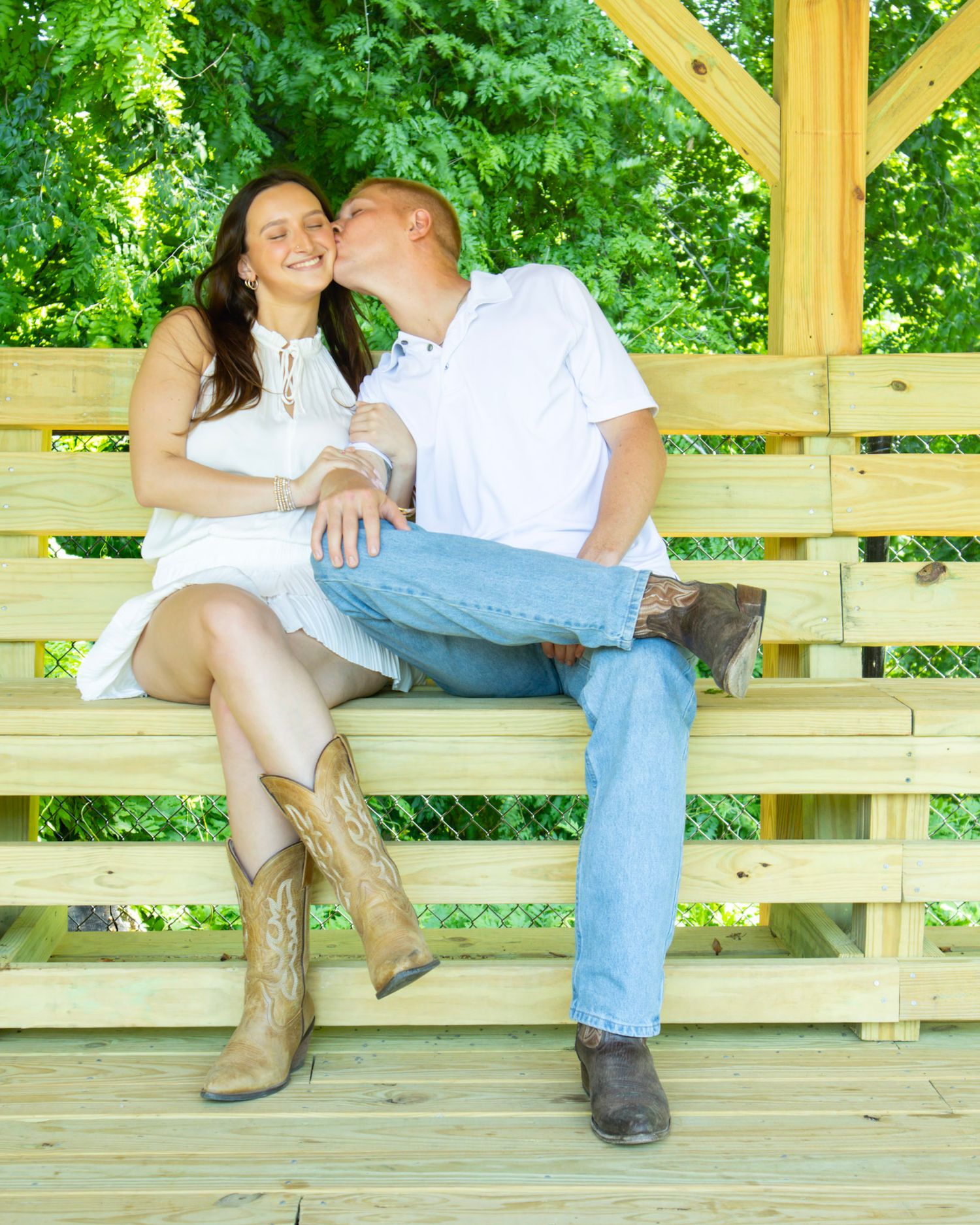 Couple sitting on wooden bench under gazebo, man kissing woman's cheek, she wears white dress and cowboy boots.