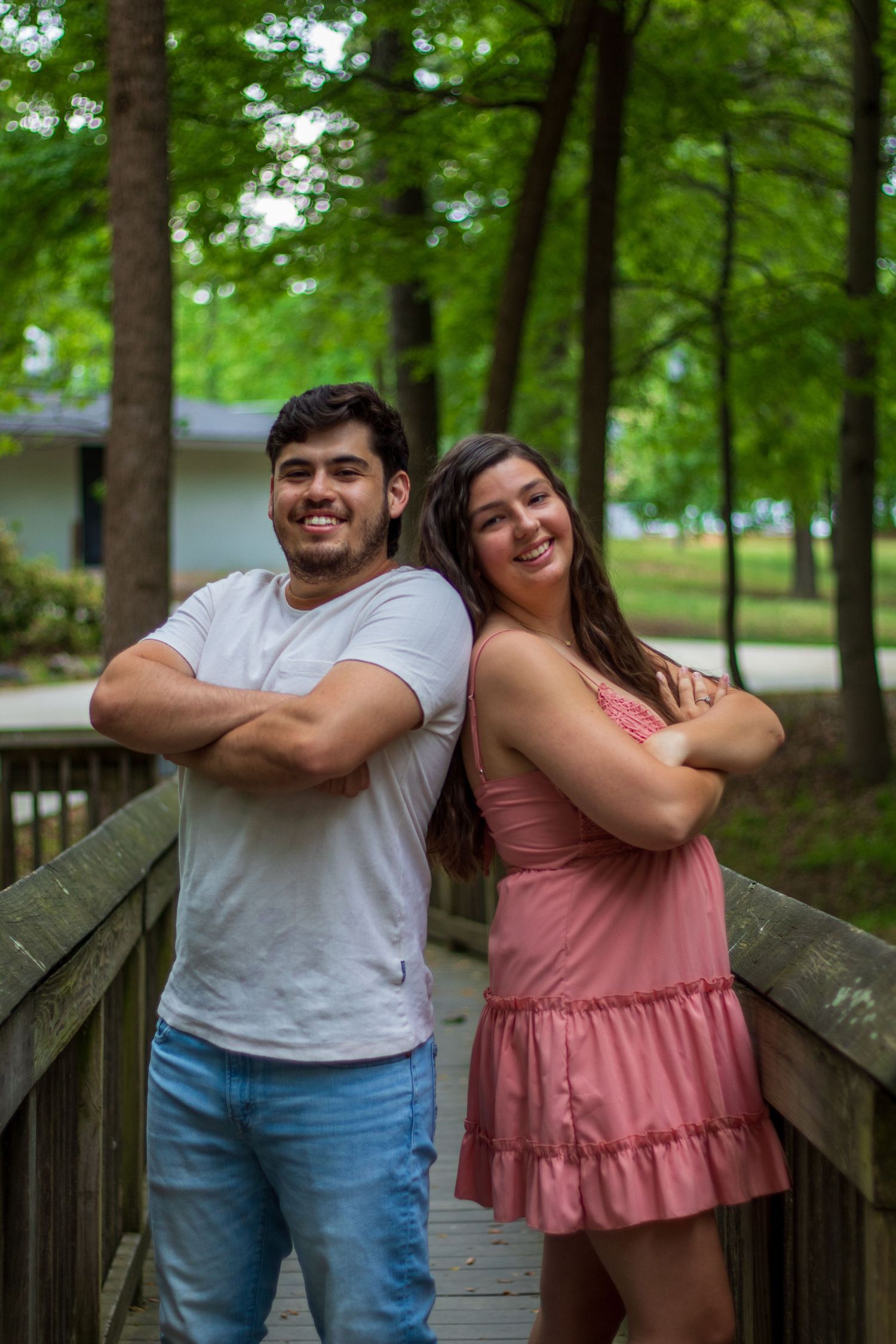 A happy couple stands back-to-back with arms crossed on a wooden bridge, smiling in a lush green park setting.