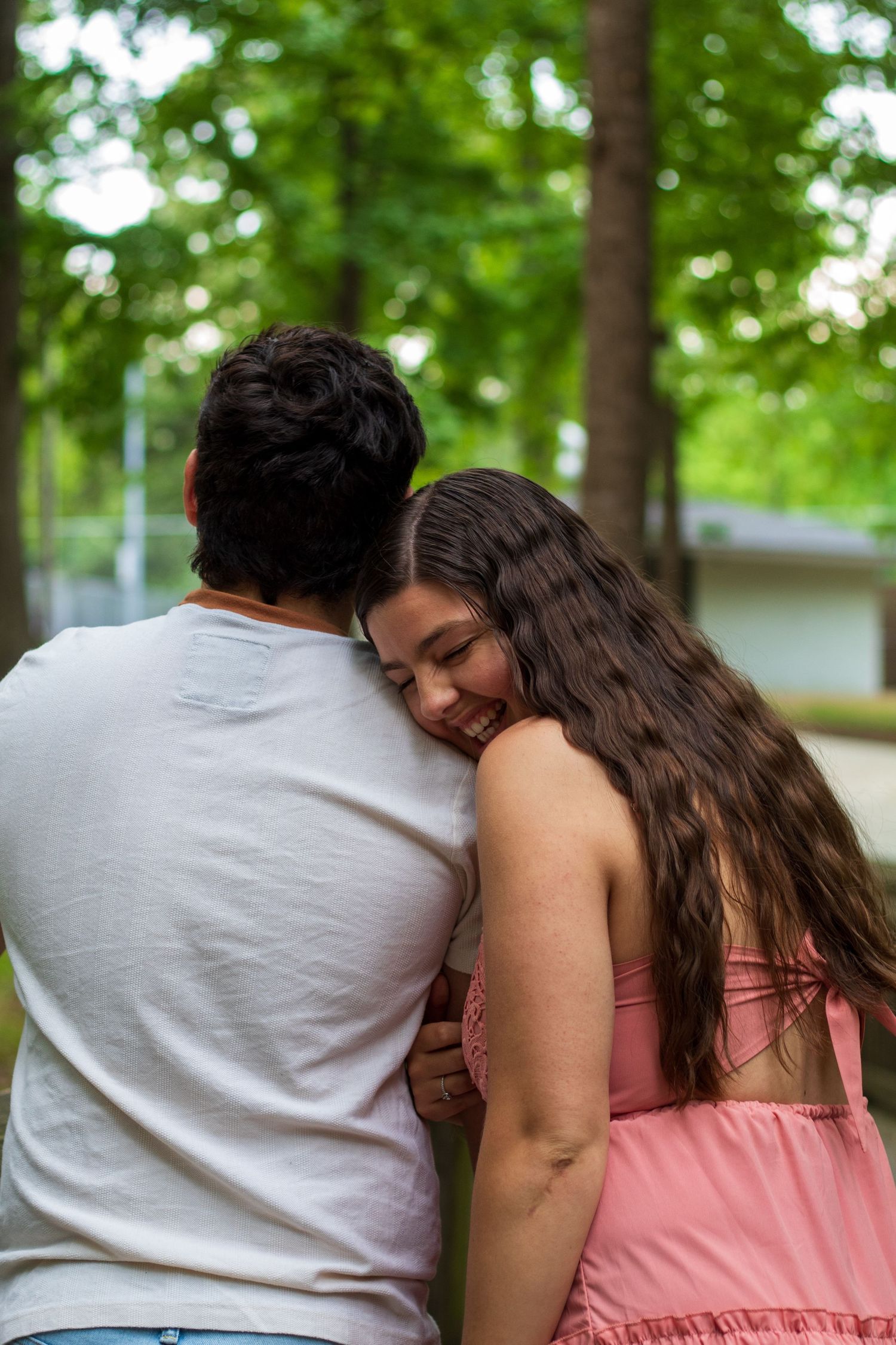Smiling woman resting head on mans shoulder from behind in green tree-lined outdoor setting.