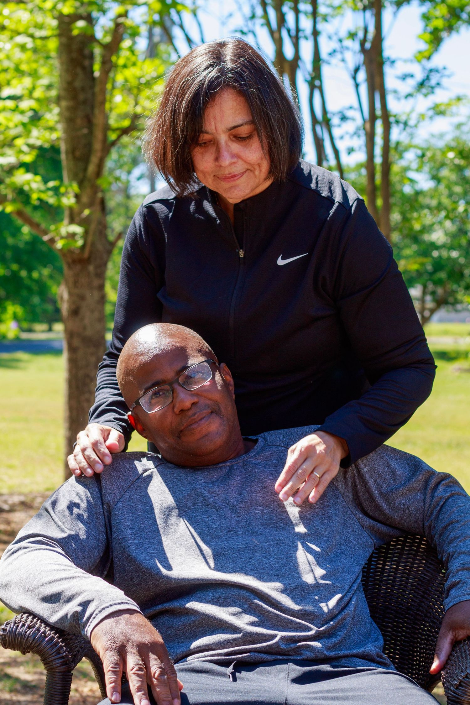 A woman in a black Nike jacket stands behind a man in a gray shirt seated outdoors in a sunny park setting.