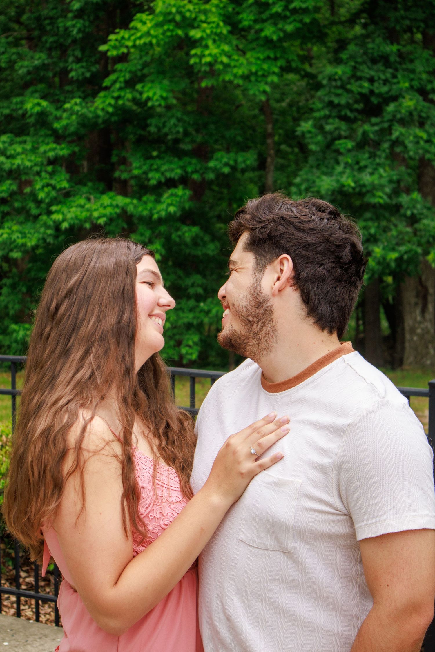 Cheerful couple gazing at each other while embracing outdoors surrounded by lush green trees.