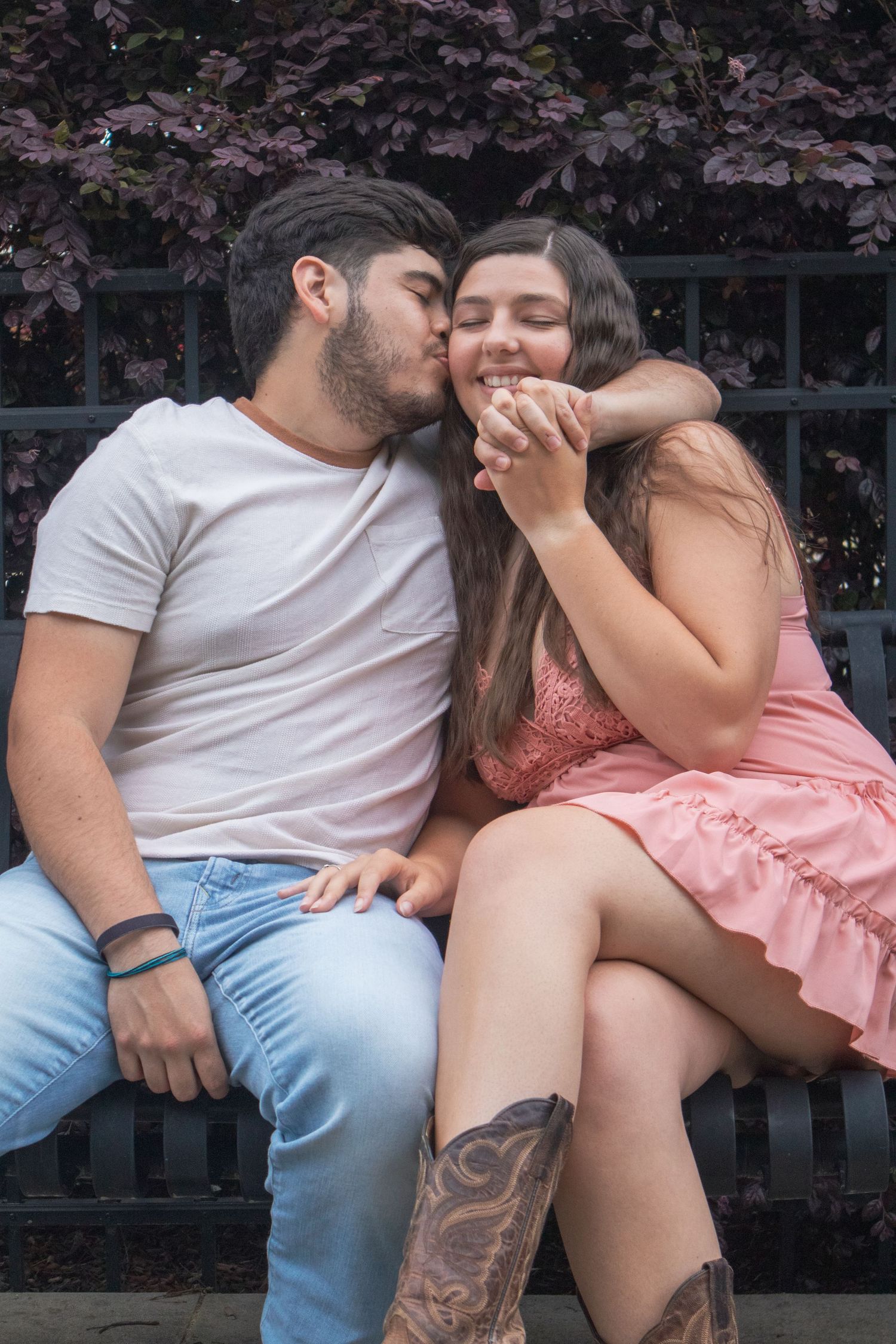 Young man kissing smiling woman on cheek while sitting on bench with purple bushes behind.