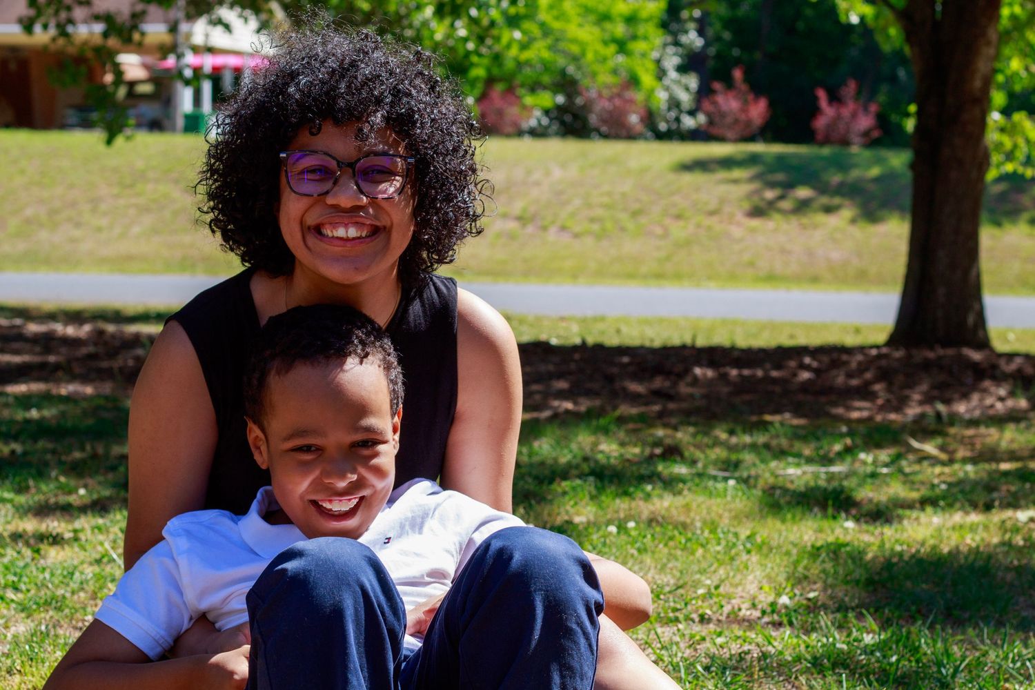 A smiling woman with curly hair and glasses sits on grass in a park with a young boy leaning against her.