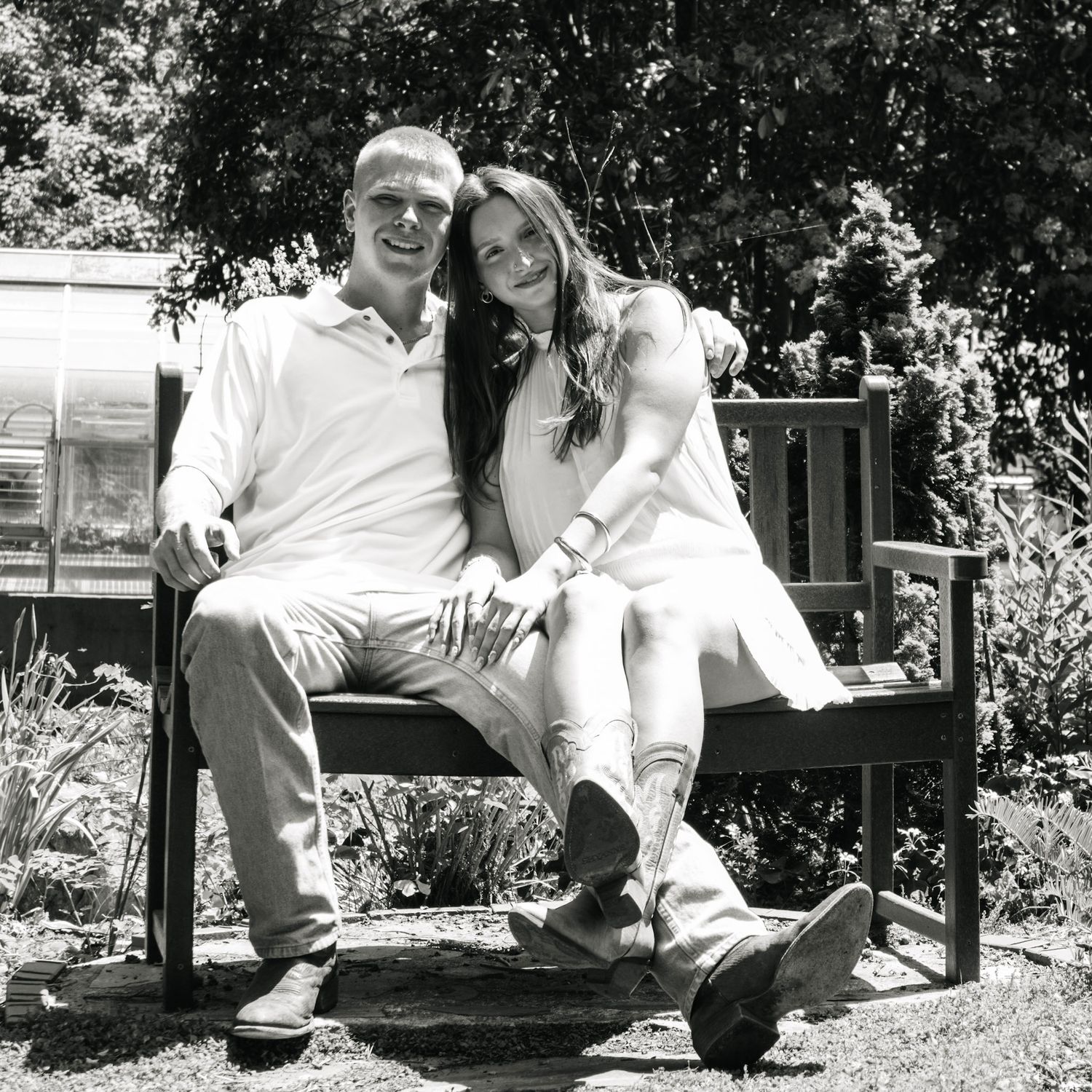 A couple smiles together on a wooden garden bench in a black and white outdoor portrait photo.
