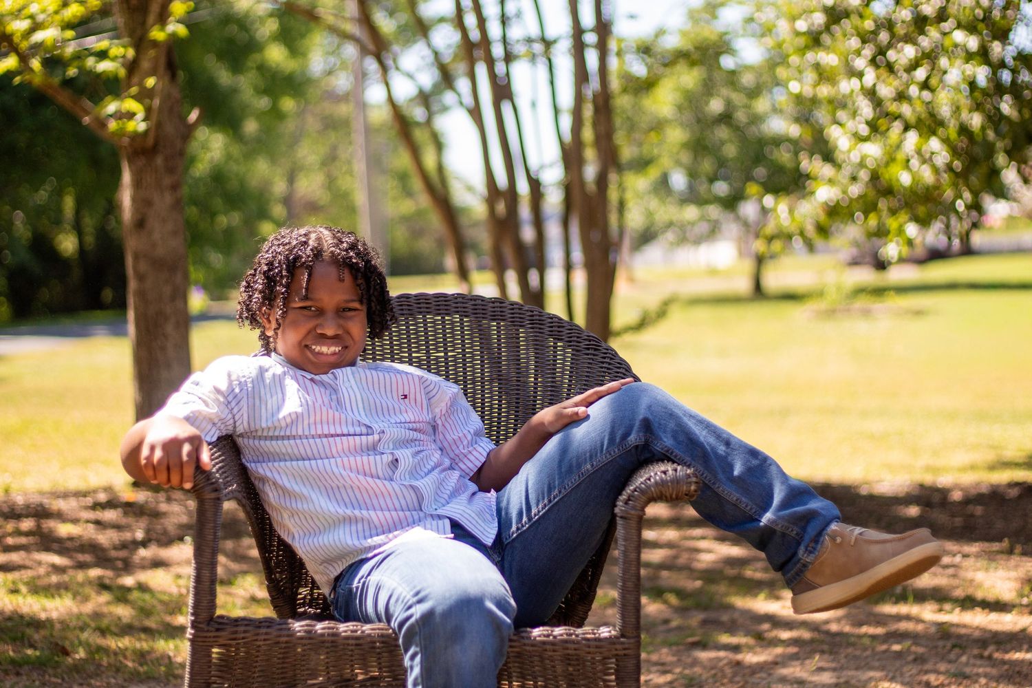 Young man smiling and relaxing in a wicker chair outdoors in a sunny park setting.