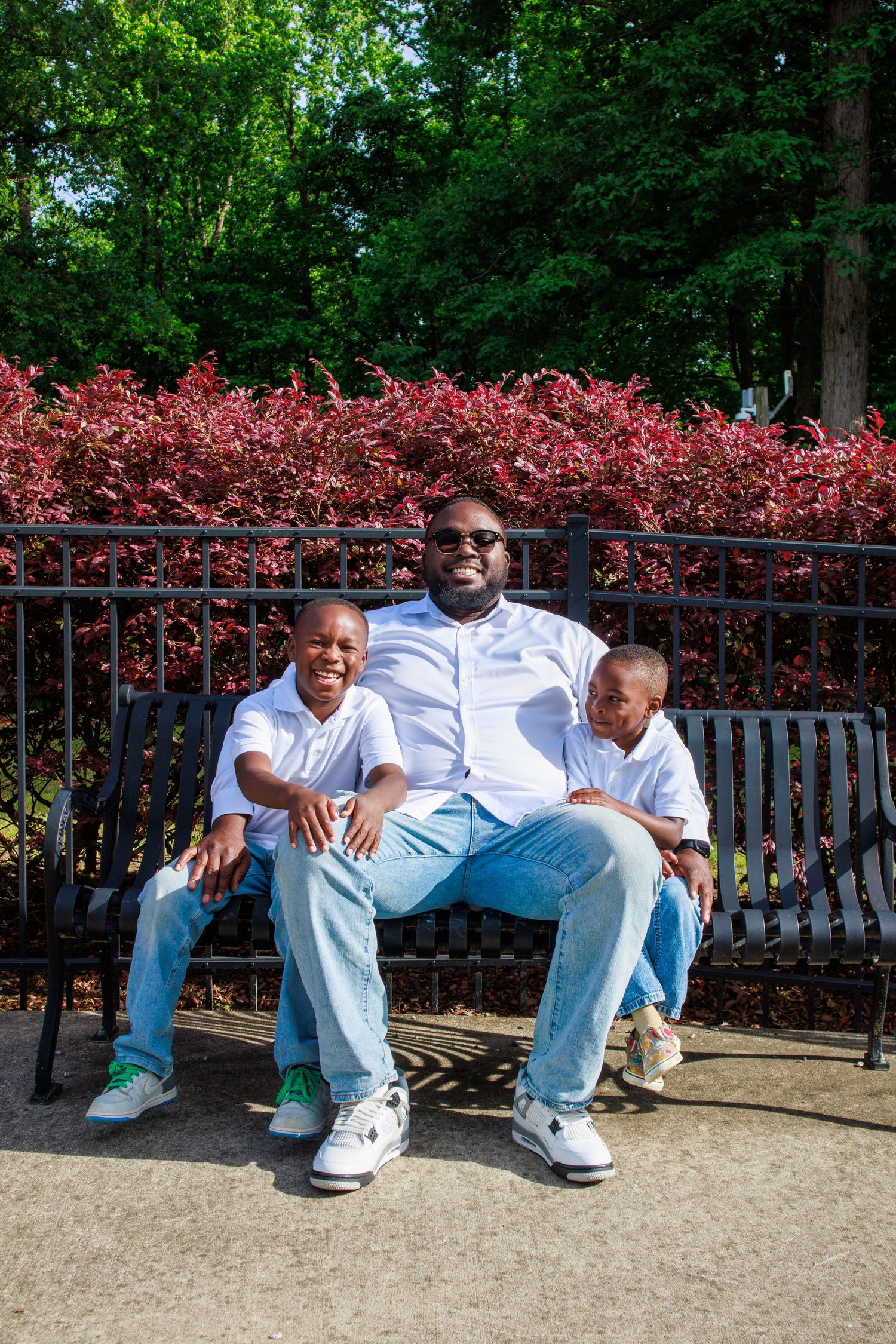 A man and two young boys sit together on a park bench, all wearing white shirts and jeans, smiling outdoors.