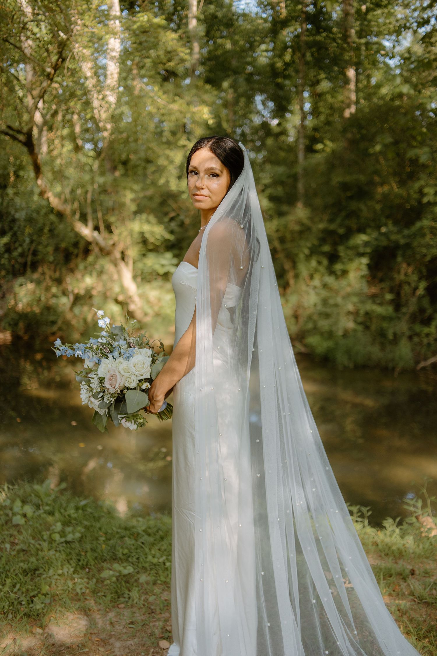 Bride in white gown with pearl veil holds blue and pink bouquet in lush green forest setting.