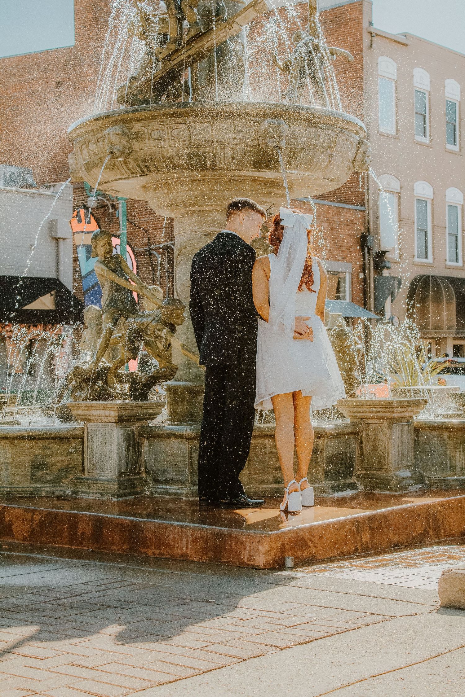 Bride and groom kiss in front of an ornate stone fountain in a charming town square.