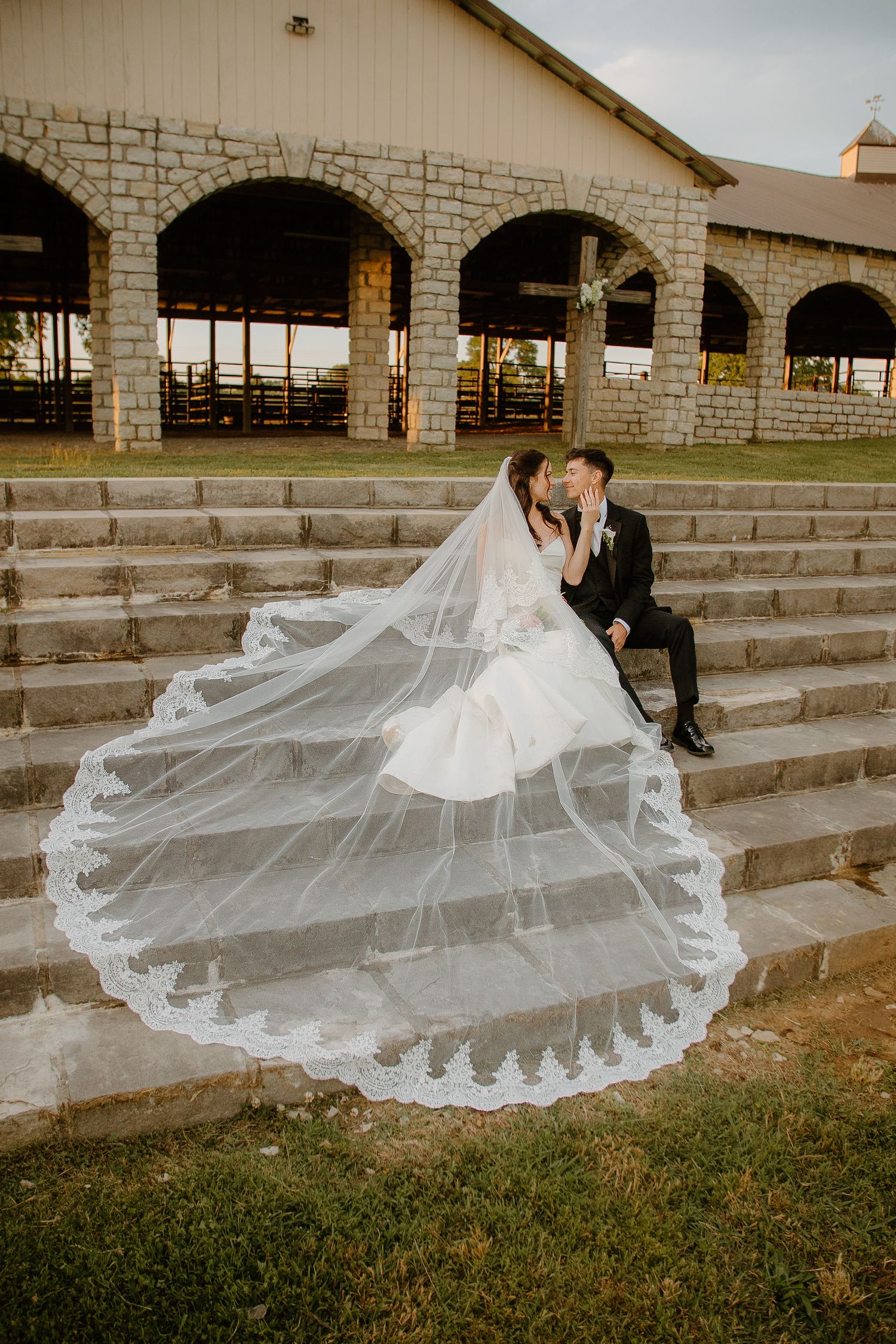Bride and groom share a romantic kiss on stone steps, her lace cathedral veil flowing dramatically, with a rustic venue behind them.
