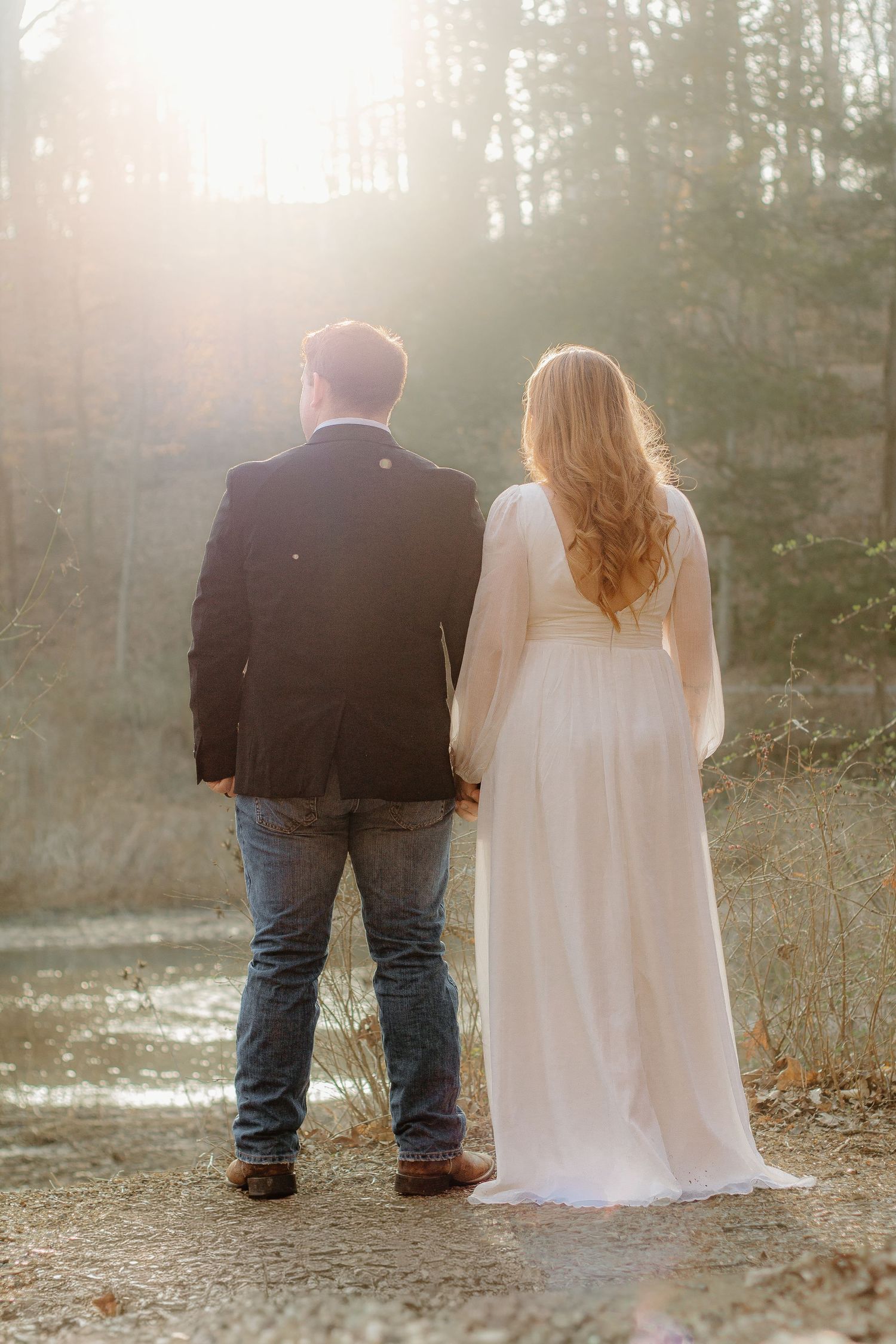 Bride in flowing white gown and groom hold hands facing a glowing sunset in a winter forest.