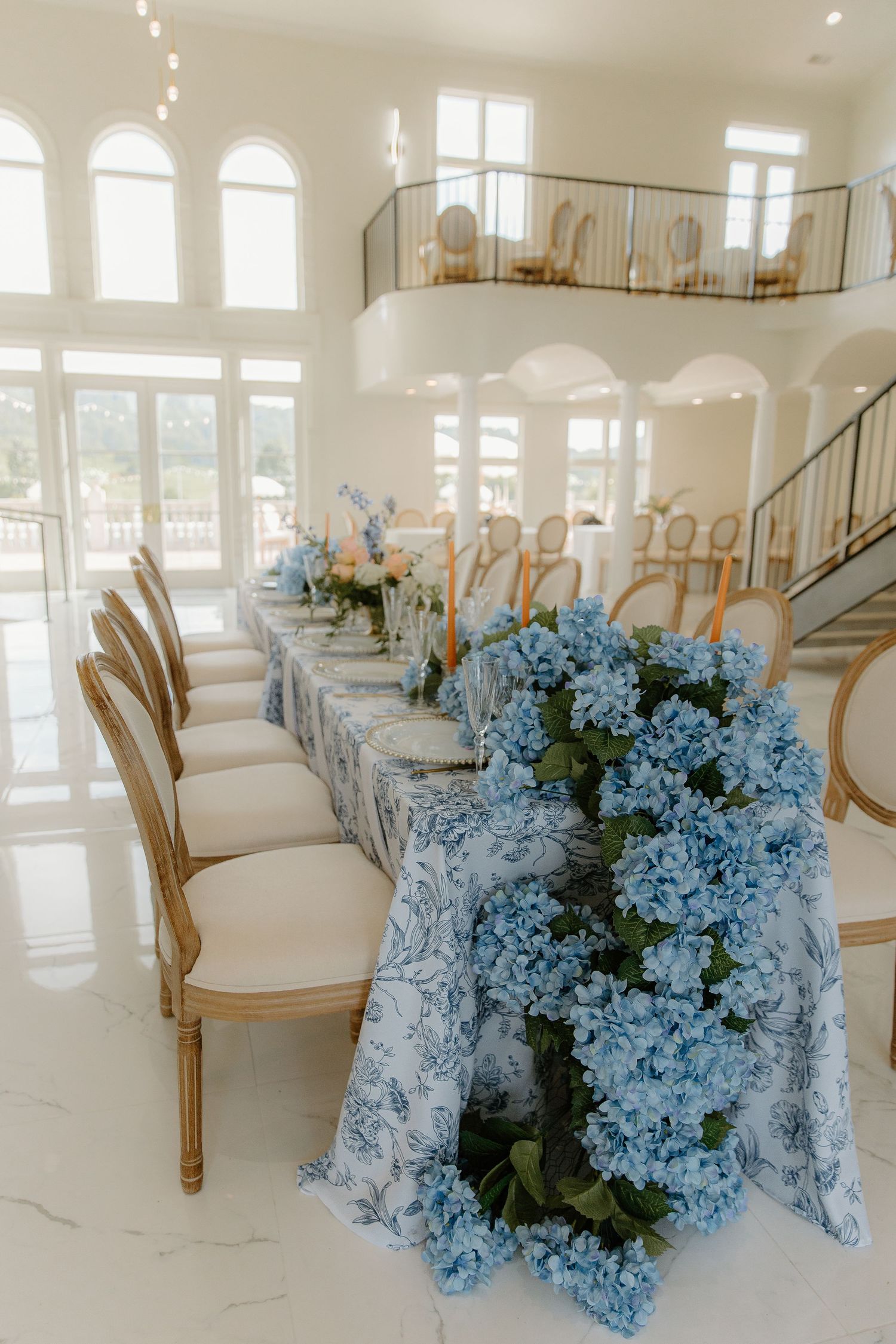 Elegant wedding reception table with blue hydrangea garland, blue floral tablecloth, gold chairs in a bright white venue.