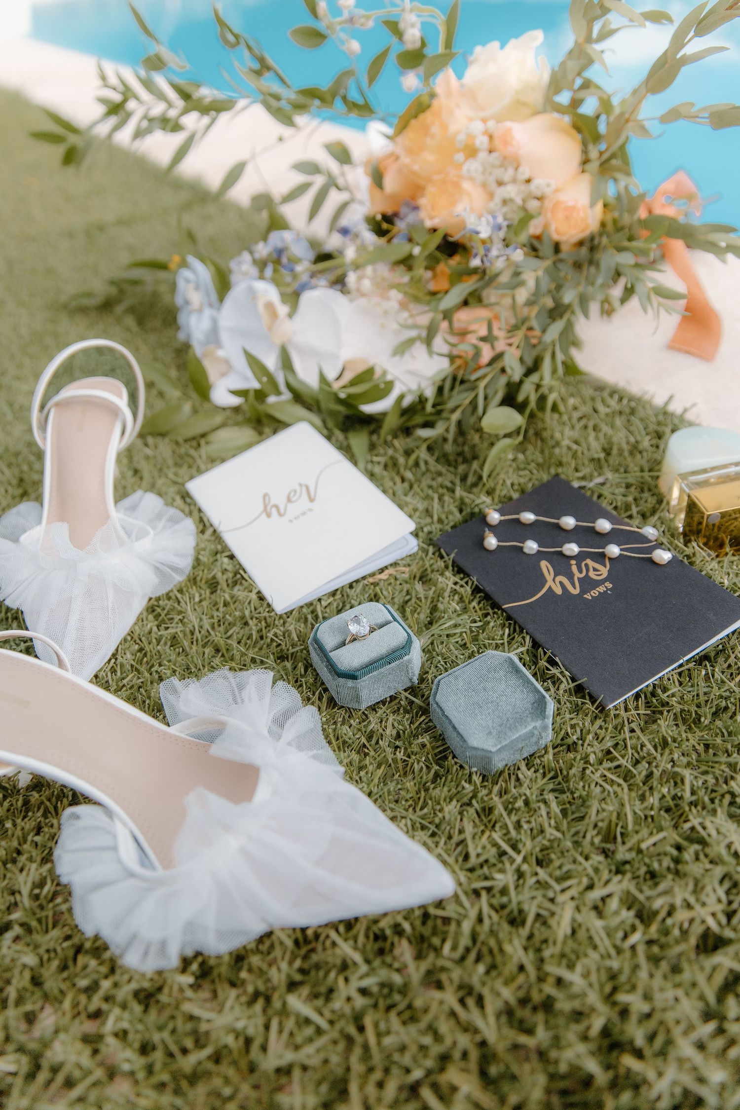 Wedding flat lay with white heels, rings, vow books, and floral bouquet on green grass.