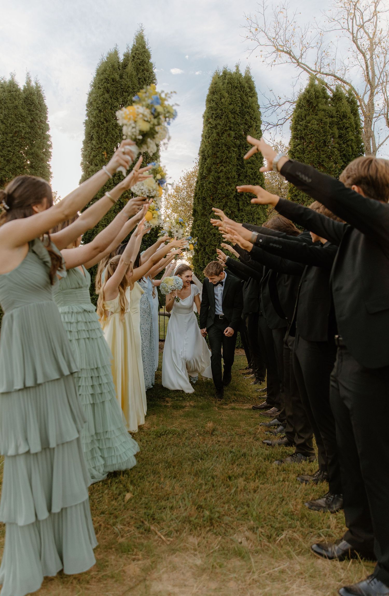 Wedding party forms tunnel arch as bride and groom walk through between cypress trees outdoors.