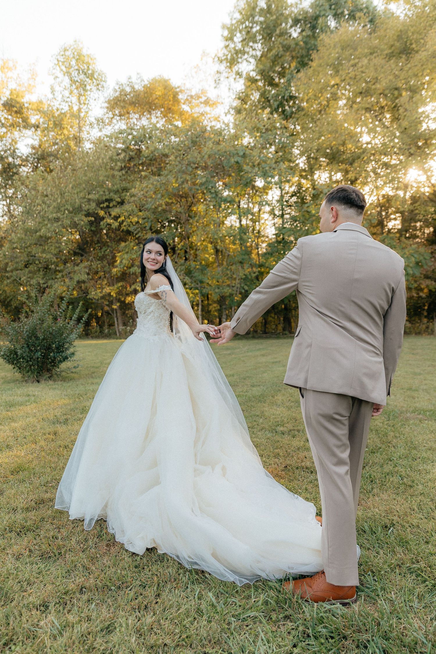Smiling bride in a voluminous white gown holds hands with groom in tan suit during golden hour outdoor shoot.