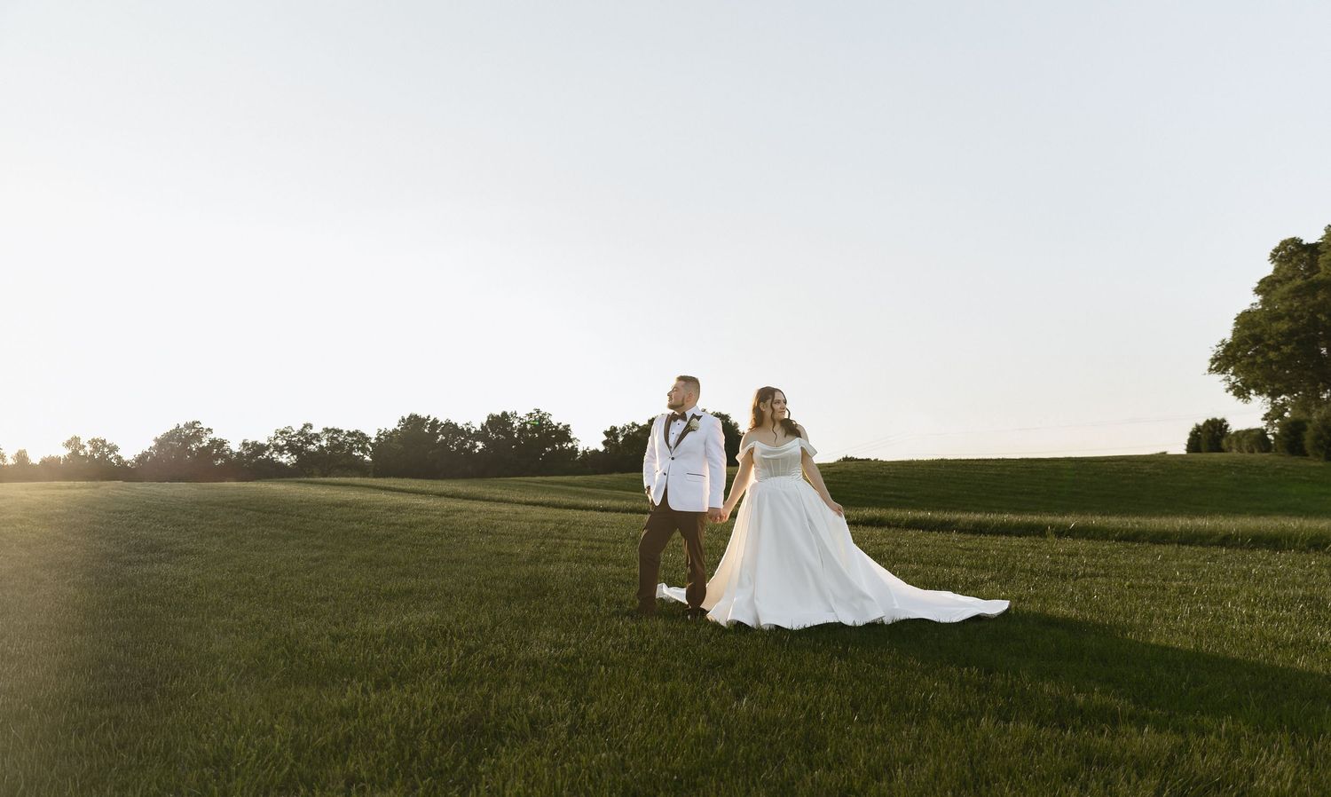 Bride and groom holding hands in a sunlit green field at golden hour, with trees in the background.