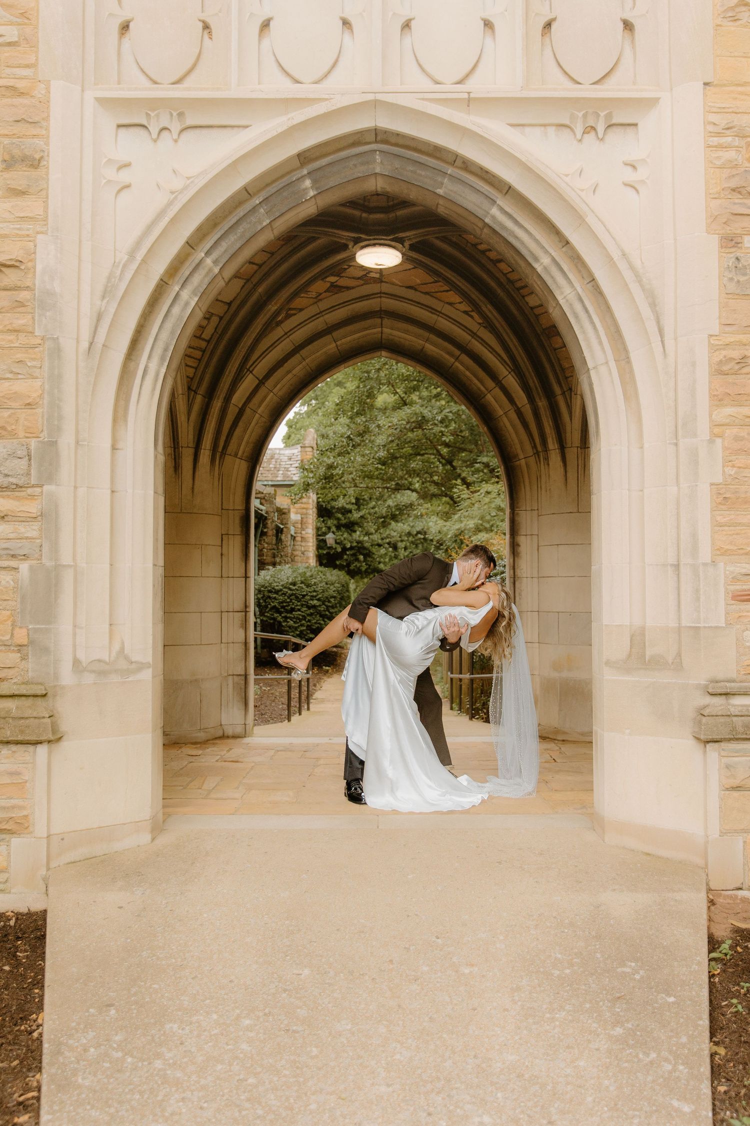 Groom dips bride under a Gothic stone archway in a romantic wedding portrait at a historic venue.
