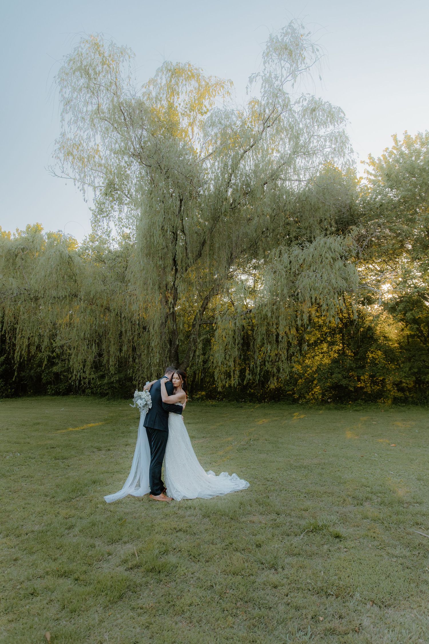 Bride and groom embracing in a green meadow with a weeping willow tree backdrop during golden hour.