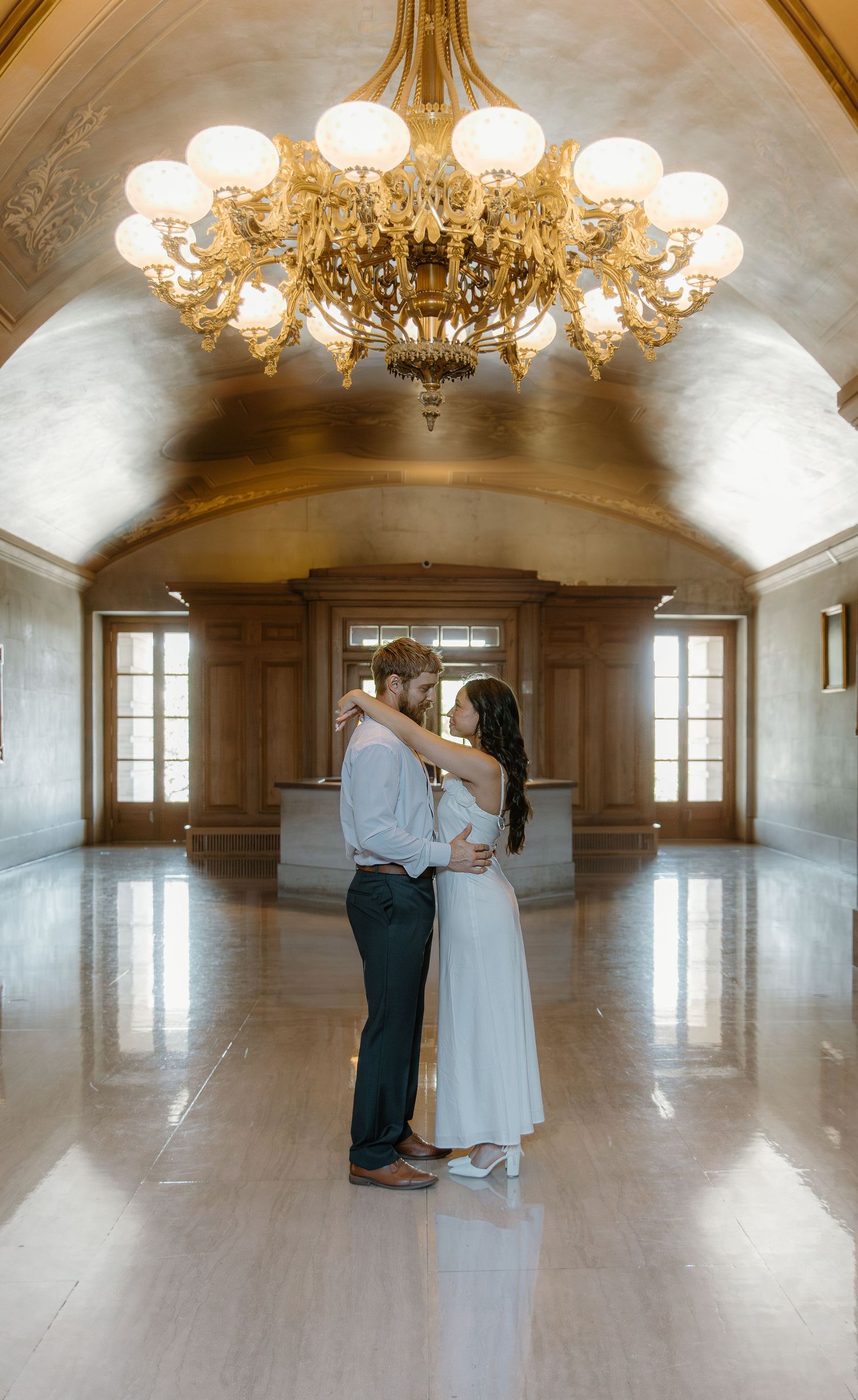 Bride and groom share a romantic embrace beneath a glowing gold chandelier in a ballroom.