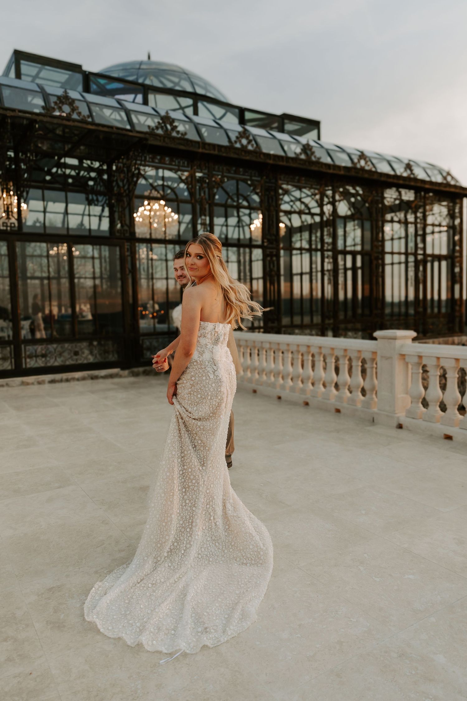 Elegant bride in a sparkling strapless gown poses on a terrace in front of a grand glass greenhouse.