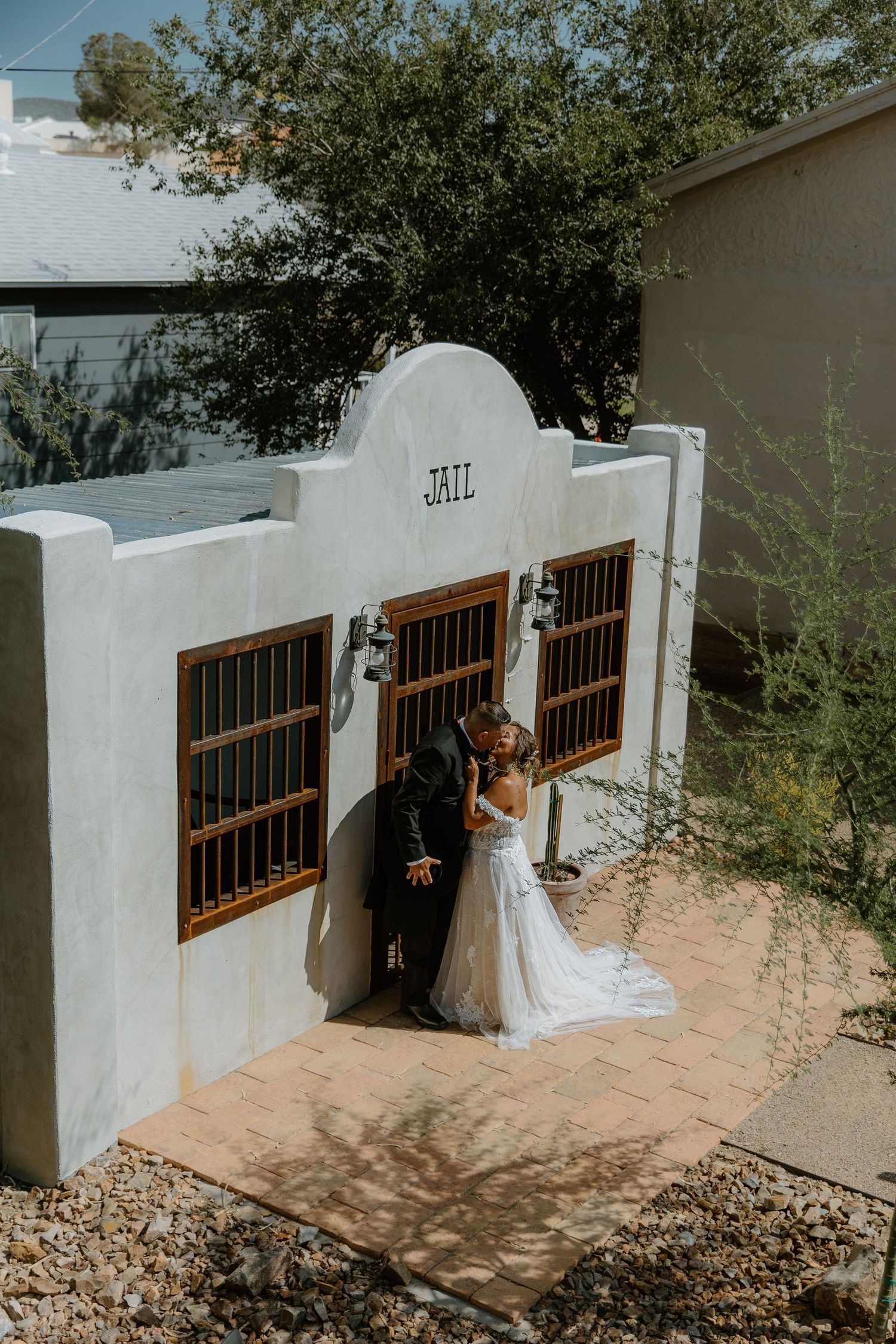 Bride and groom share a kiss in front of a rustic white adobe jail building at an outdoor wedding venue.