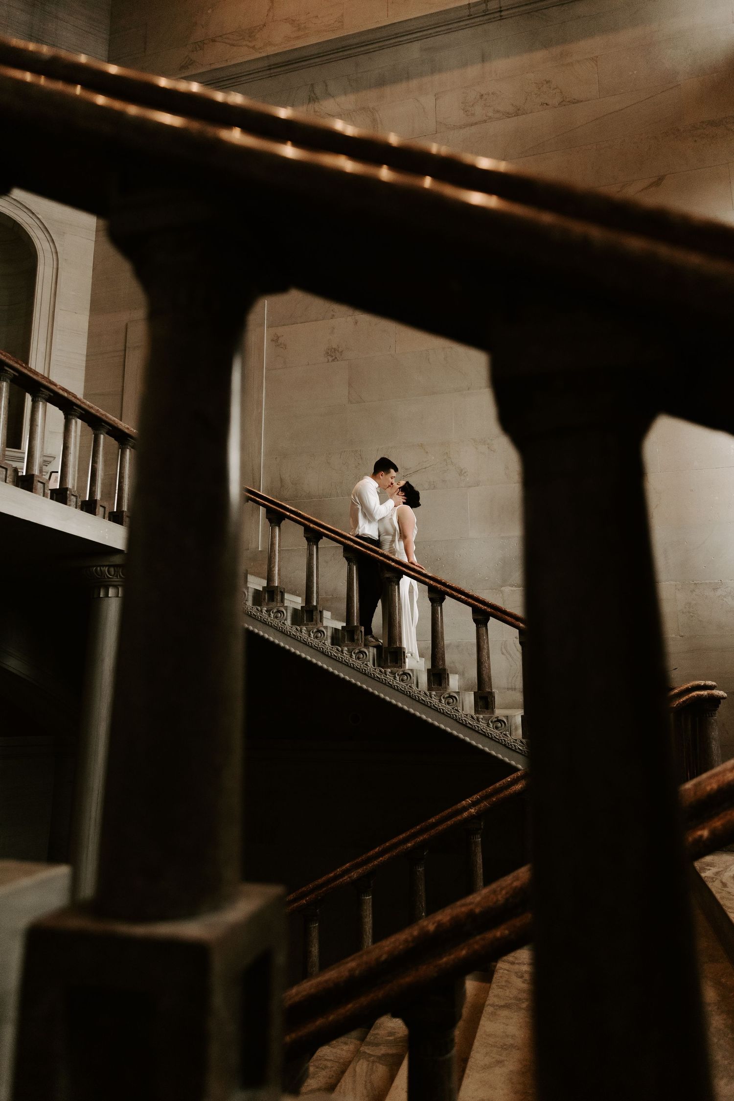 Couple shares a romantic kiss on an ornate wooden staircase in a grand historic building interior.