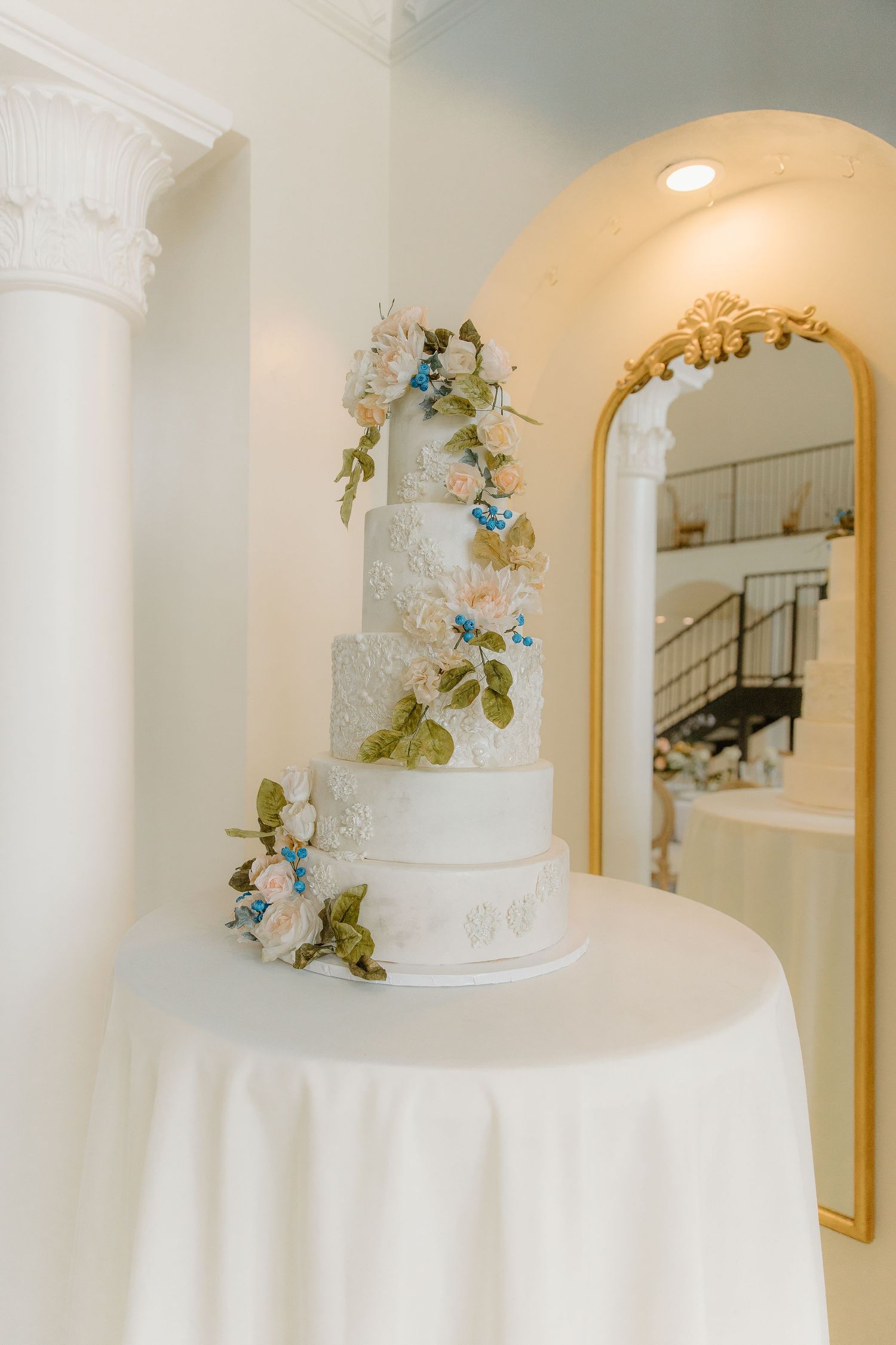 Elegant five-tier white wedding cake adorned with pink and blue sugar flowers displayed on a draped table.