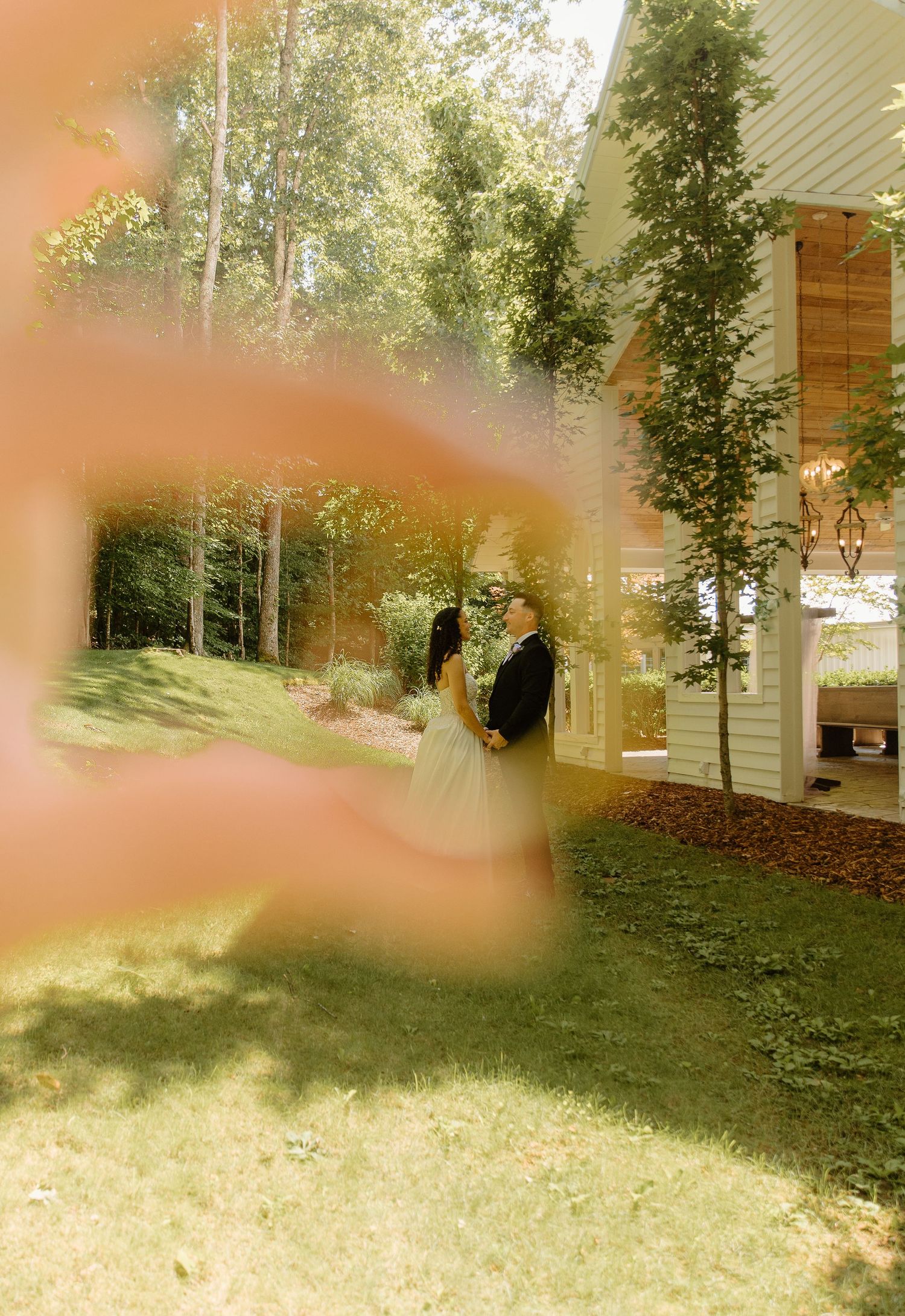 A bride and groom stand together outdoors near a white building, framed by a blurred pink foreground and lush green trees.