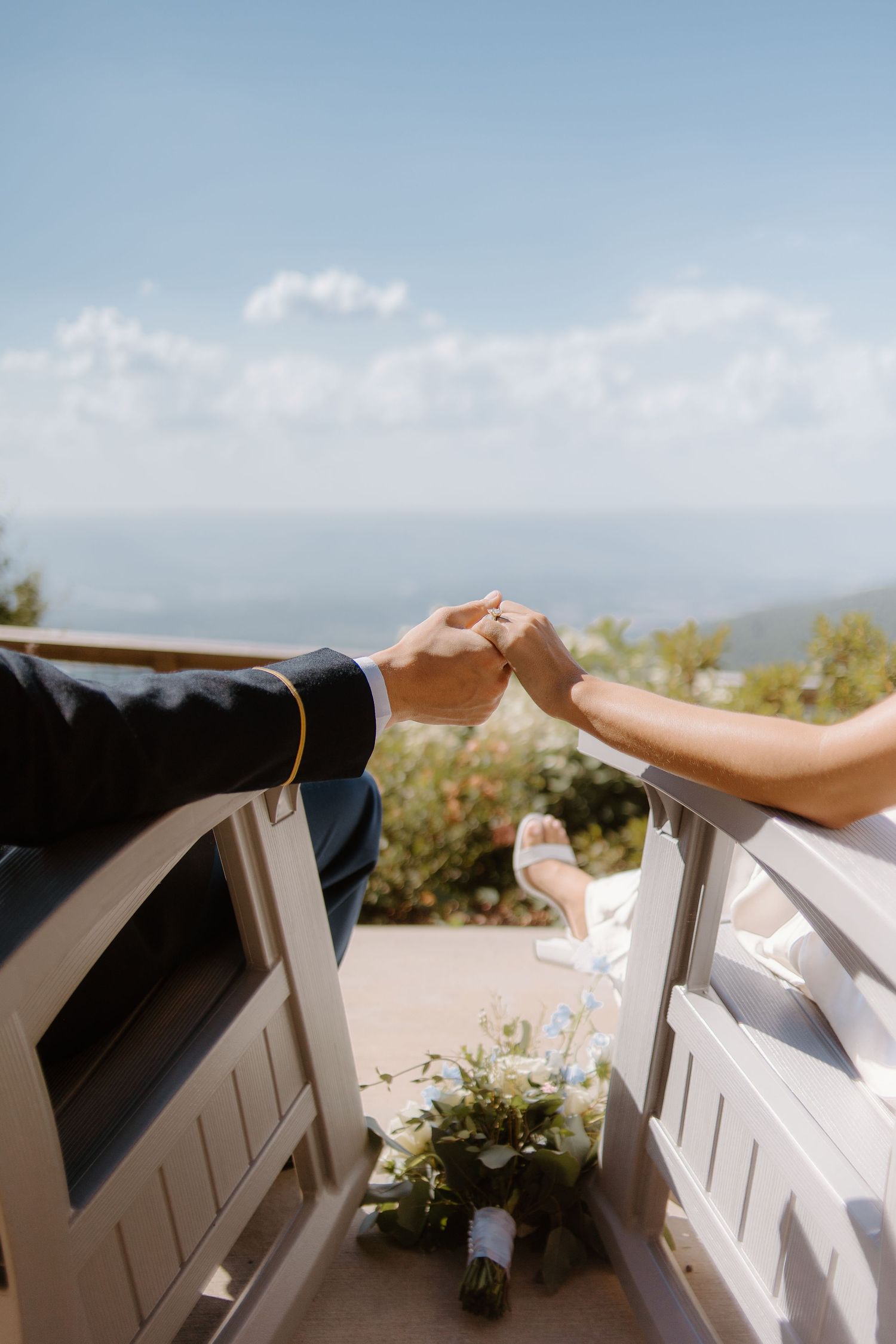 Newlyweds holding hands on white benches with ocean view and bouquet.