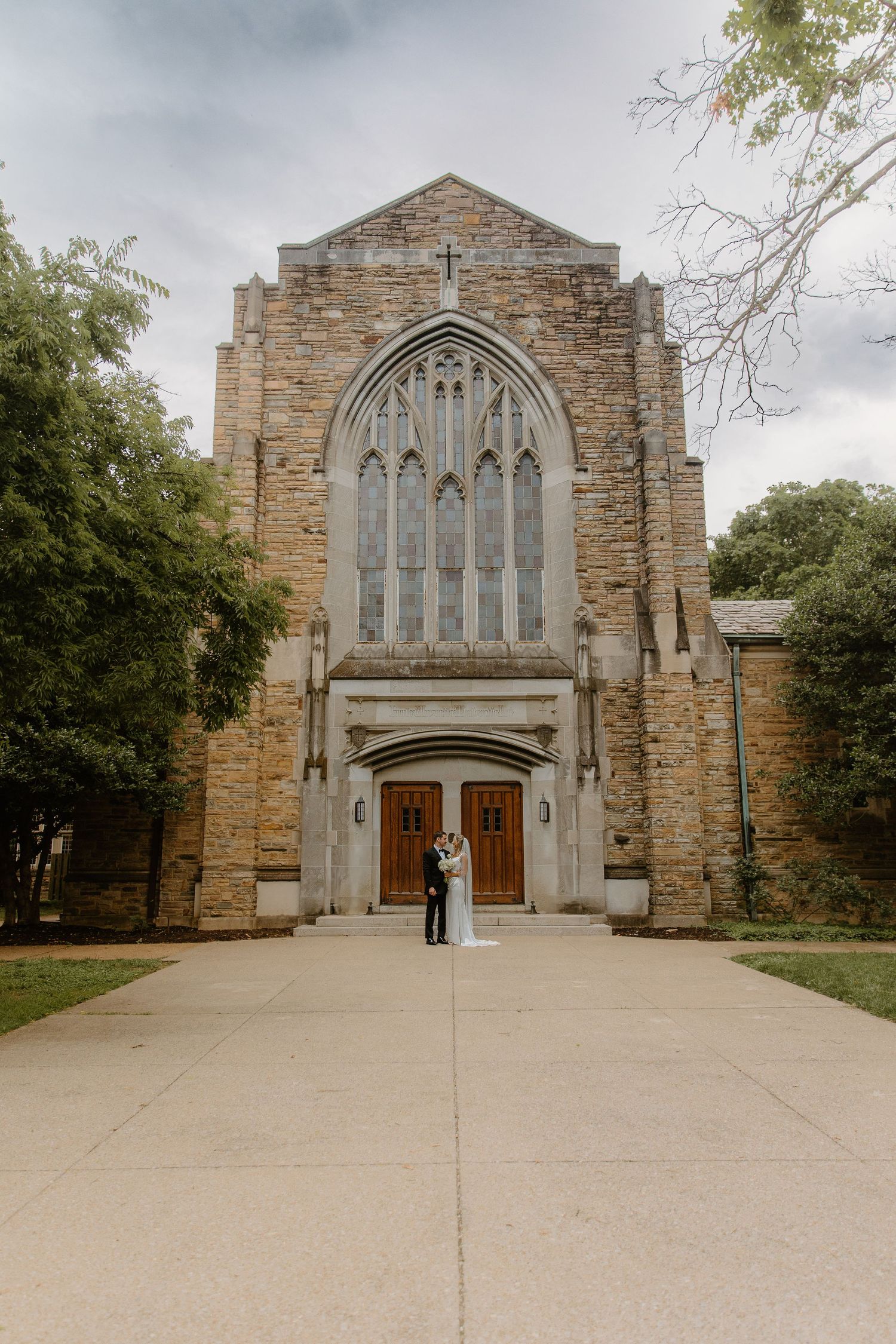 Wedding couple kissing before grand Gothic stone church with large arched stained glass window.